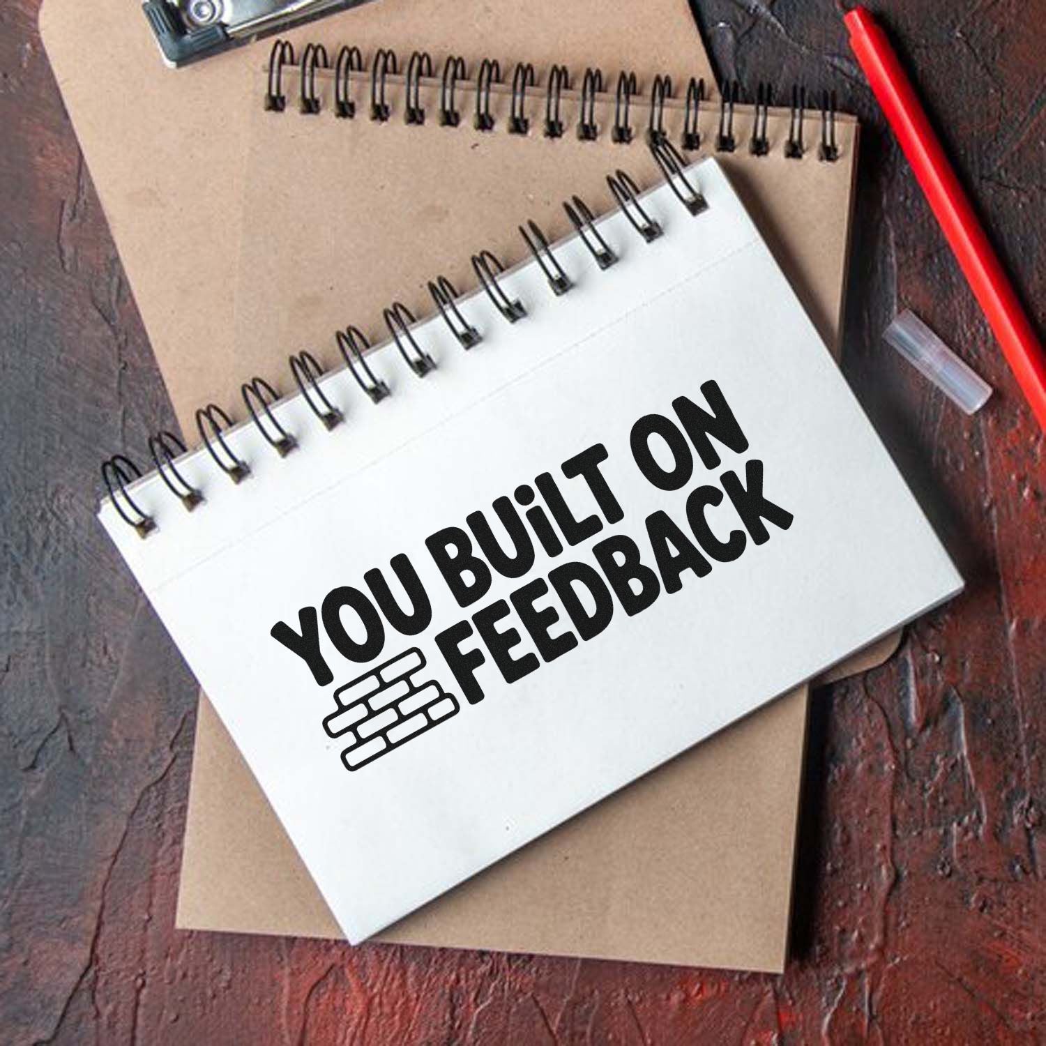 White spiral notebook with black stamp-style You Built on Feedback seal and brick icon, on kraft notebook beside a red pen.
