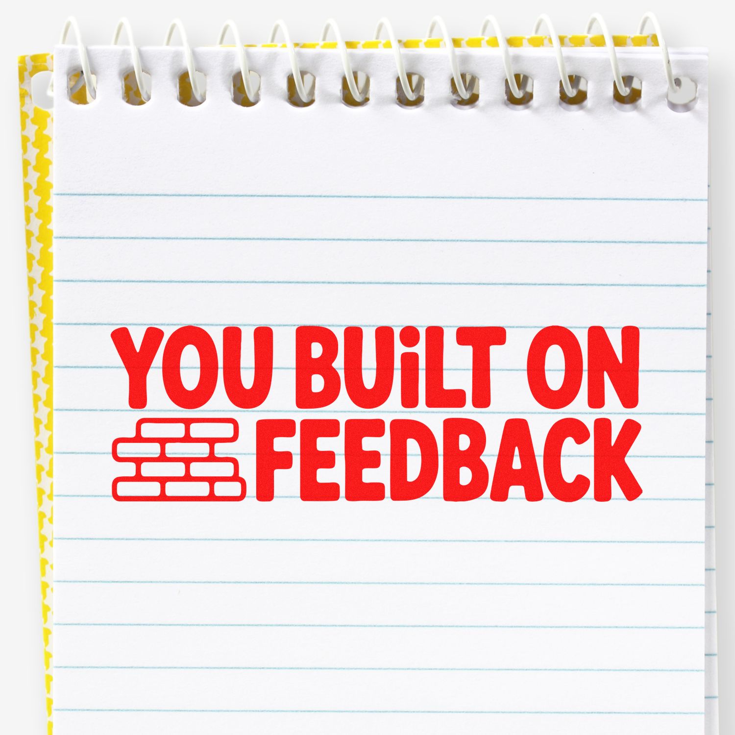 Red feedback stamp on white spiral notepad with brick icon, blue lines, and yellow patterned border.