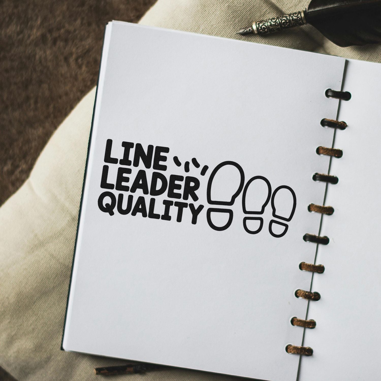Black Line Leader Quality stamp seal with footprint icon on white spiral notebook page, brown binding, and pen.