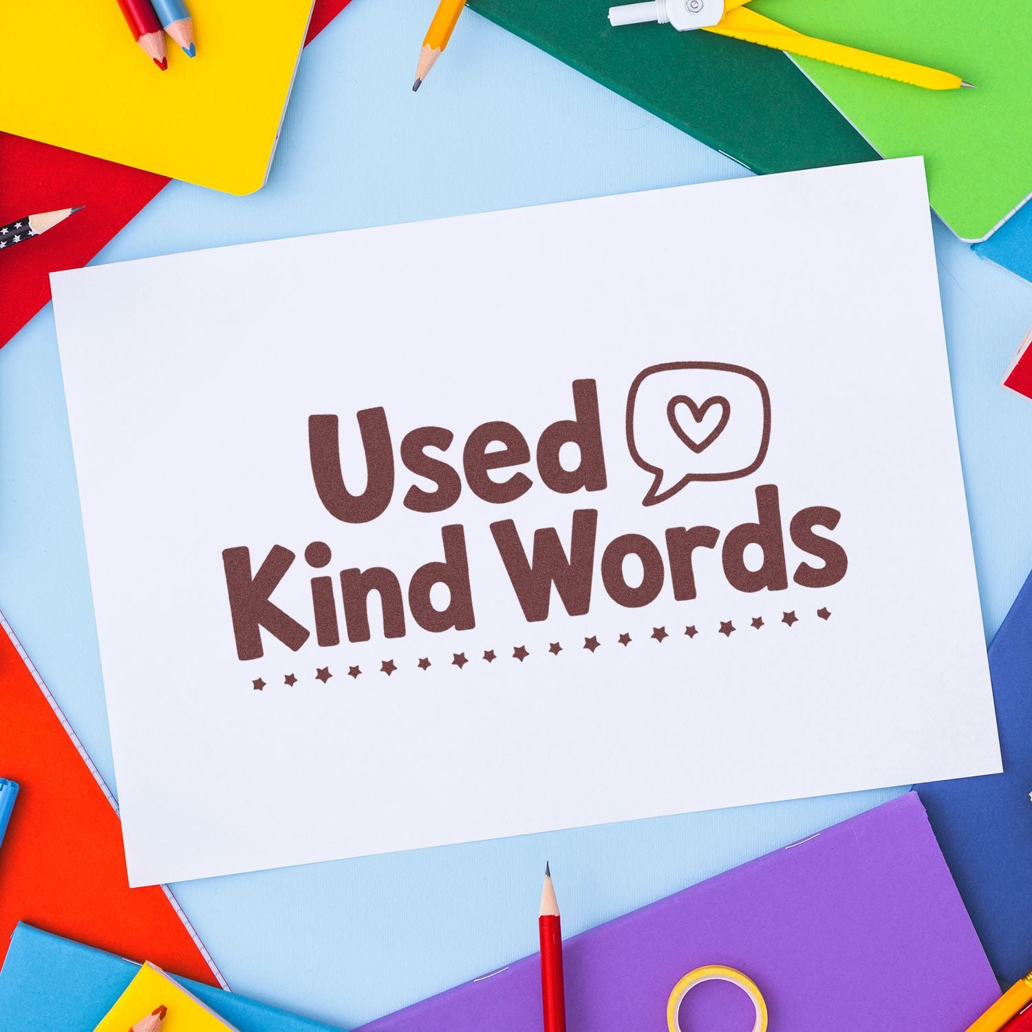 Brown Used Kind Words stamp with heart seal on white card, surrounded by colorful pencils and notebooks.