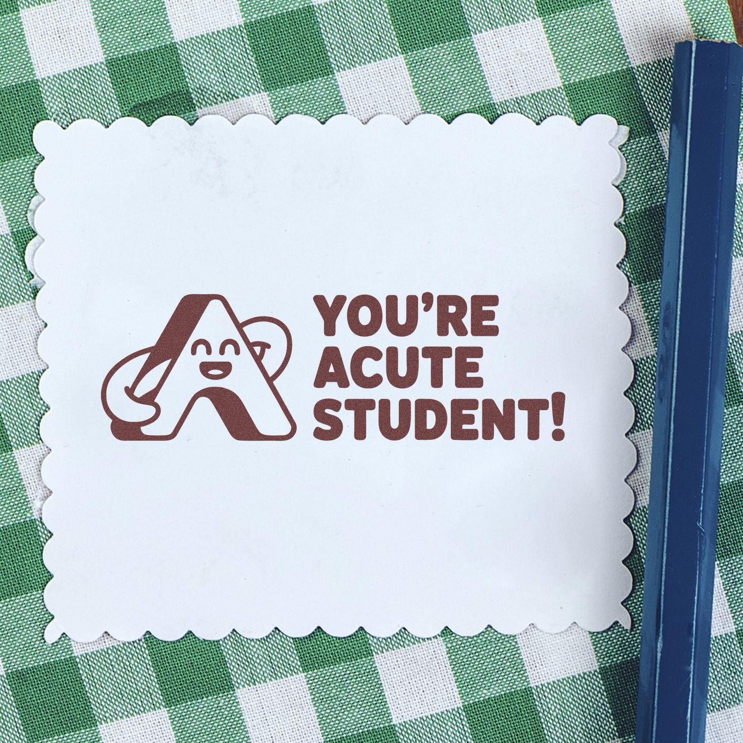 White scalloped stamp seal with brown Youre acute student! text and logo on green gingham fabric, blue pen at right.
