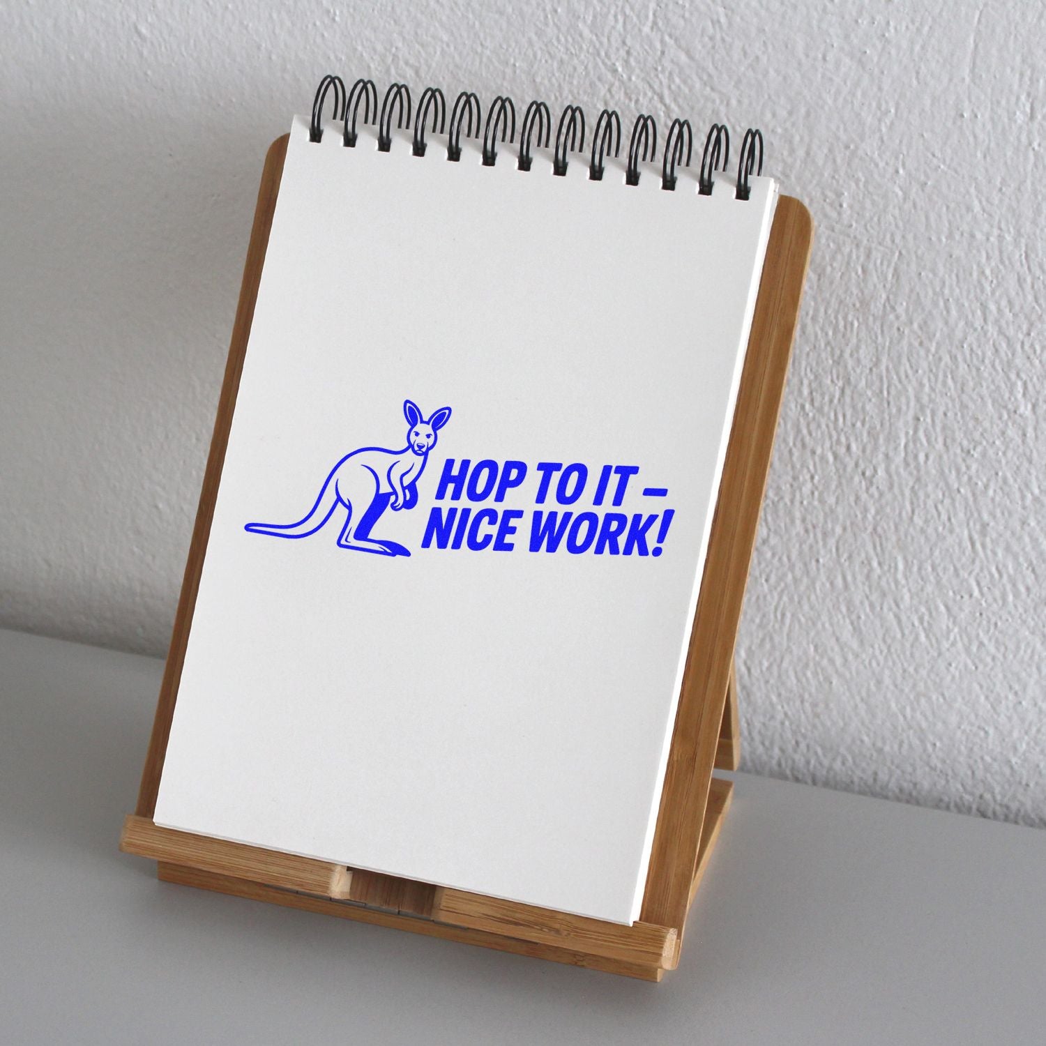 Blue kangaroo rubber stamp reading Hop to It Nice Work! on a white notepad in a wooden stand.
