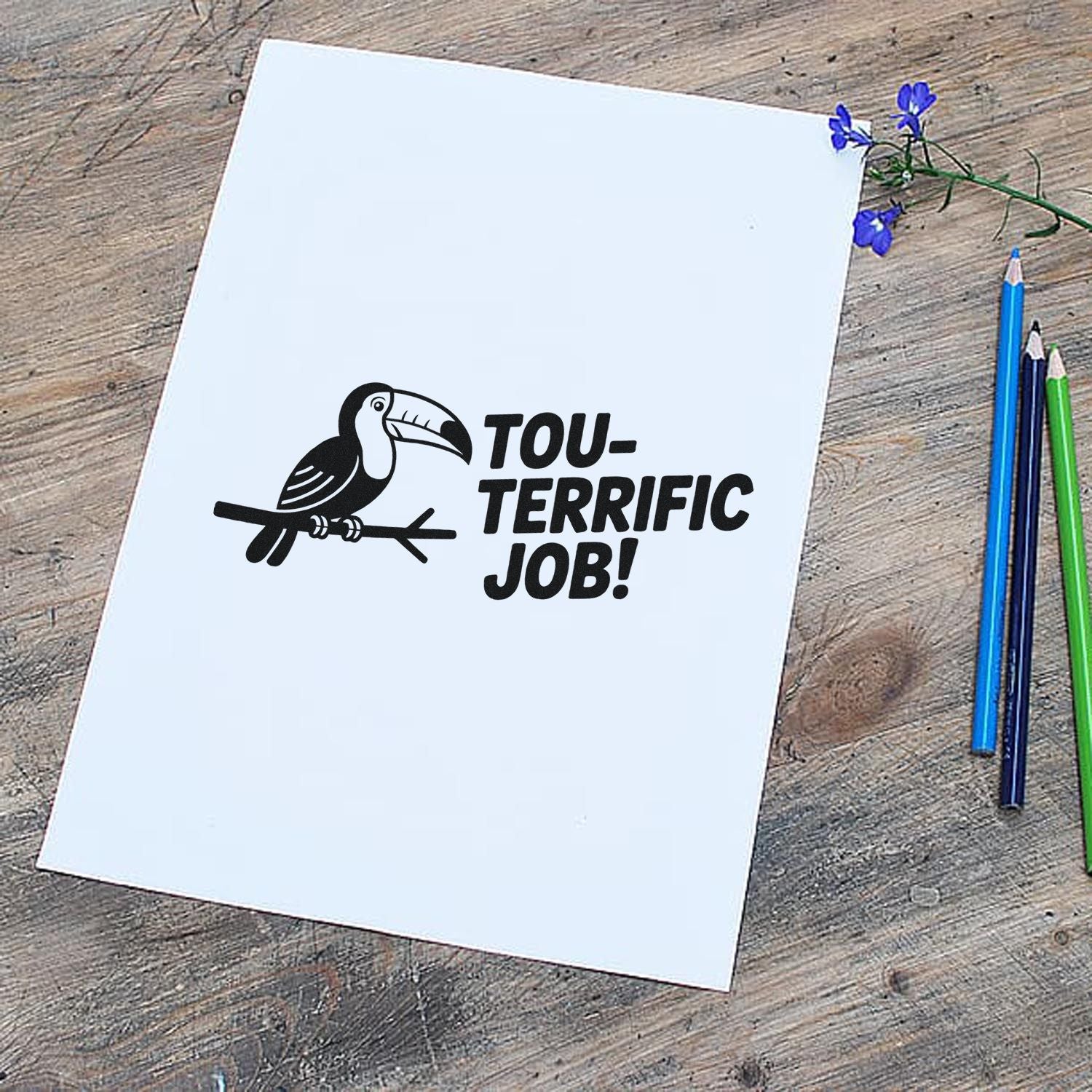 Black toucan rubber stamp seal with Tou-terrific Job! on white paper, beside blue, green and purple pencils.