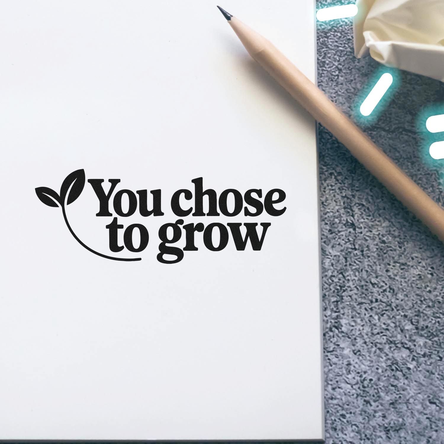 Black custom stamp and seal reading You chose to grow with leaf logo on white paper, beside a tan pencil on gray surface.