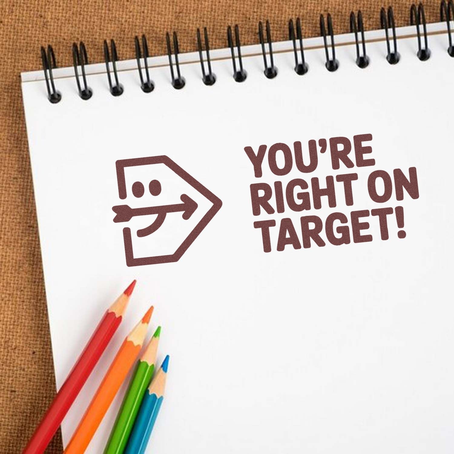 Brown stamp seal with arrow icon and YOURE RIGHT ON TARGET! on a white spiral notebook, with colored pencils.