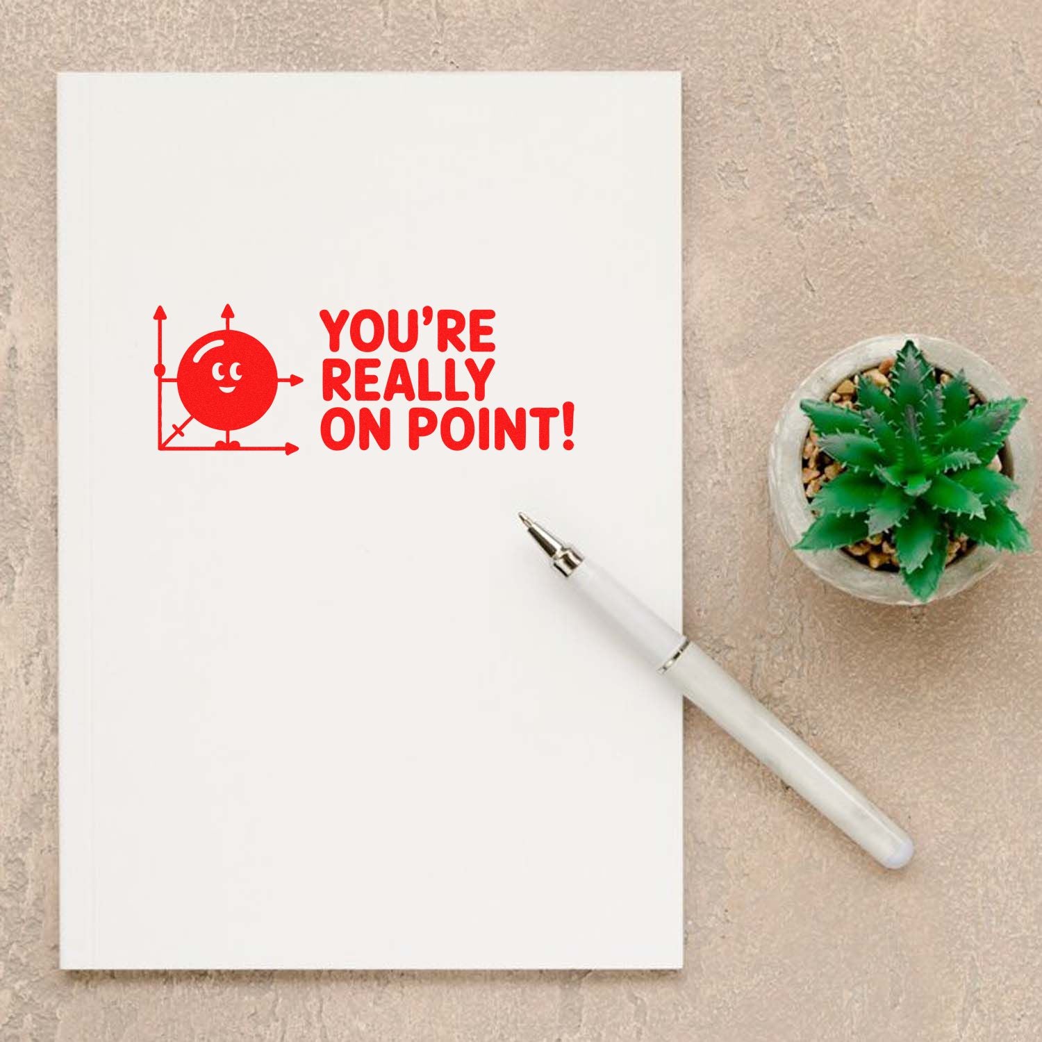 White notebook with red stamp-and-seal design saying YOURE REALLY ON POINT! beside a white pen and green succulent.