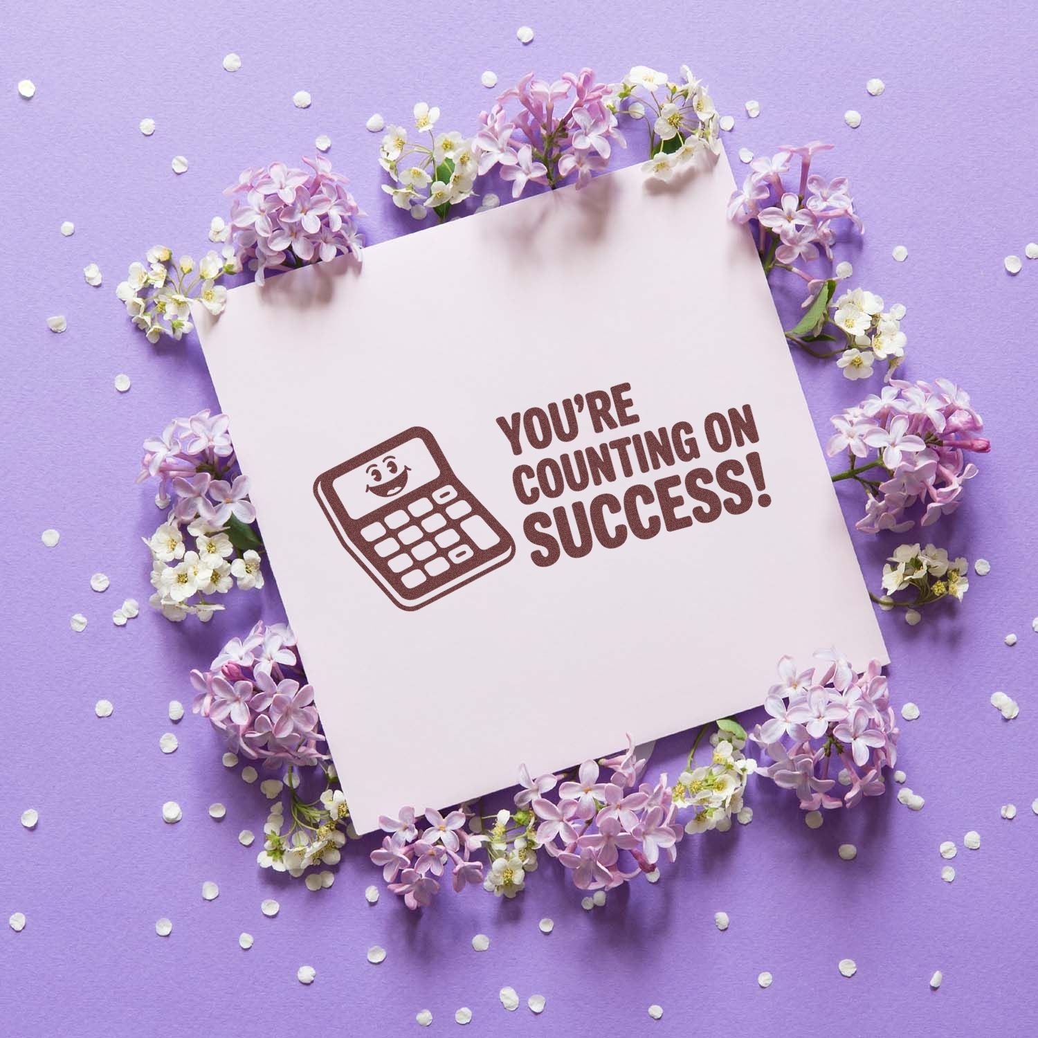 Brown calculator stamp and seal with Youre counting on success! on a white card, framed by lilac flowers on a lavender background.