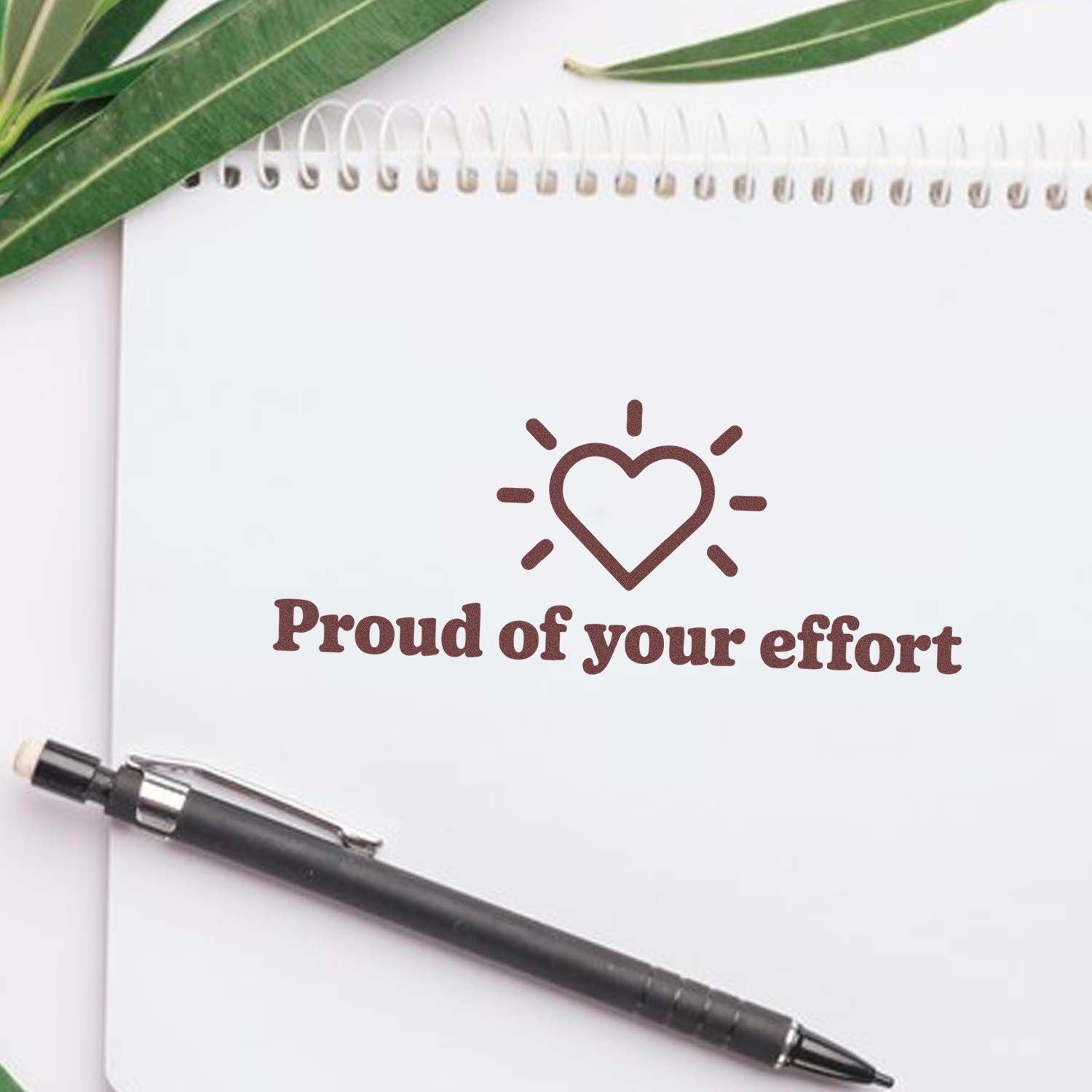 Brown heart stamp seal reading Proud of your effort on white spiral notebook, with black pen and green leaves.