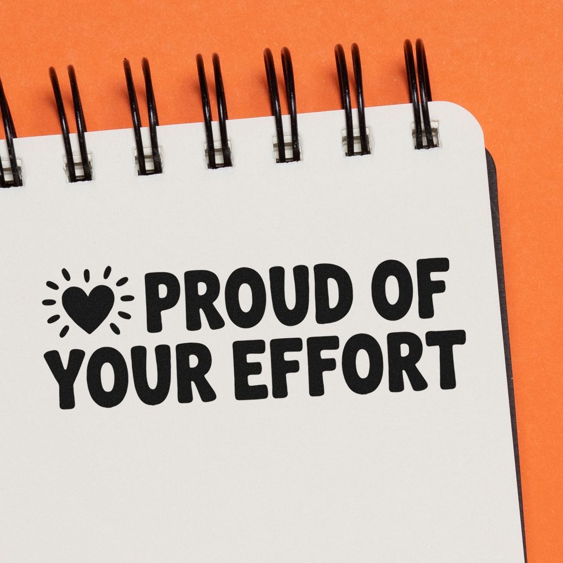 Black custom stamp and seal on a white spiral notebook reads Proud of Your Effort with a heart icon on orange background.