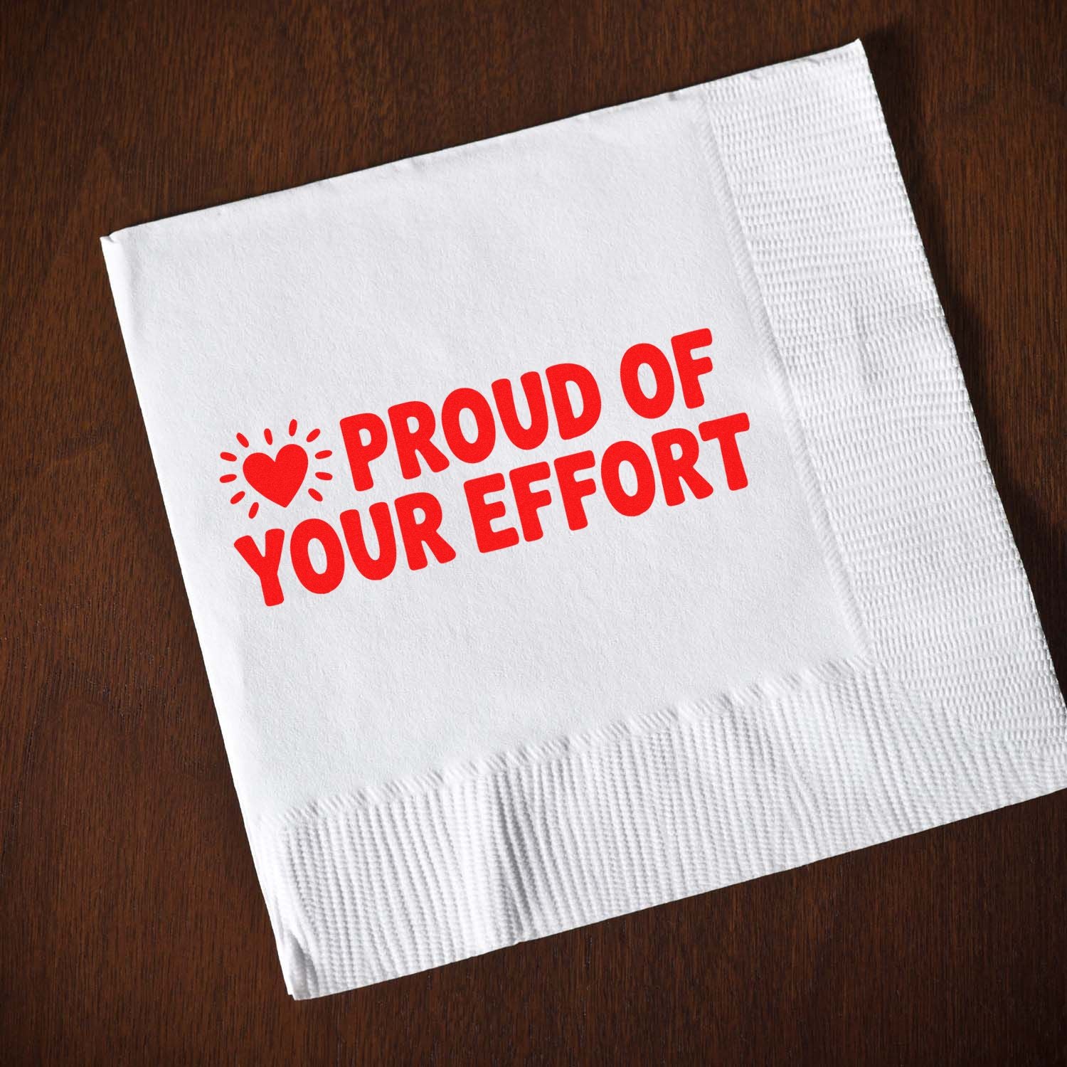 White napkin with red Proud of Your Effort stamp and heart seal on dark wood background.
