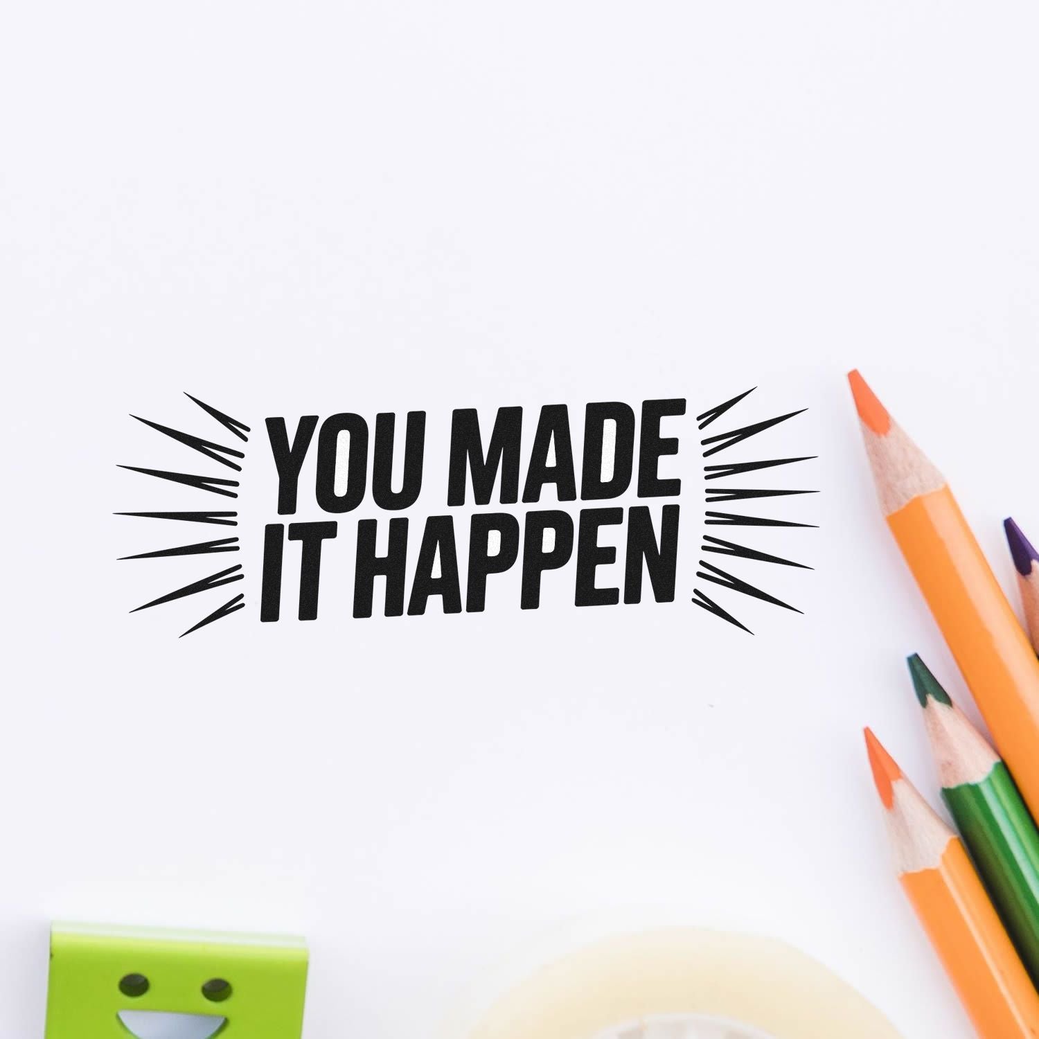 Black YOU MADE IT HAPPEN motivational stamp/seal with burst lines on white paper, surrounded by orange, green, and purple pencils.