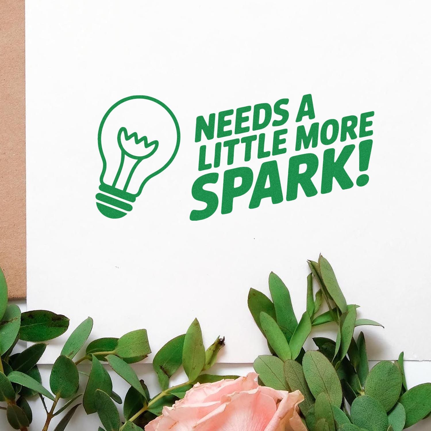 Green custom stamp seal with lightbulb icon and Needs a little more spark! text on white paper, framed by eucalyptus and pink rose.
