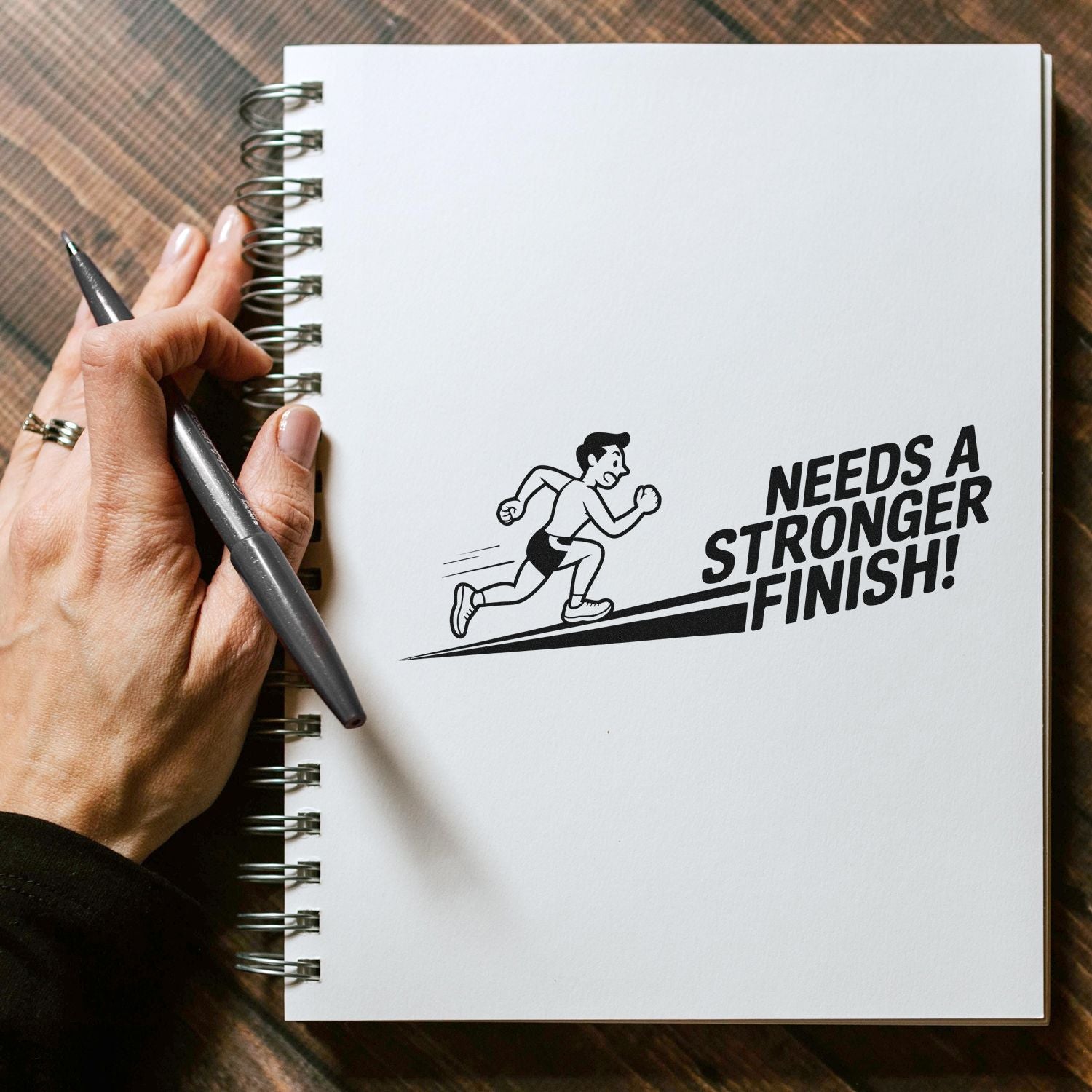 Black stamp seal on a white spiral notebook, showing a running man and bold Needs a Stronger Finish! text.