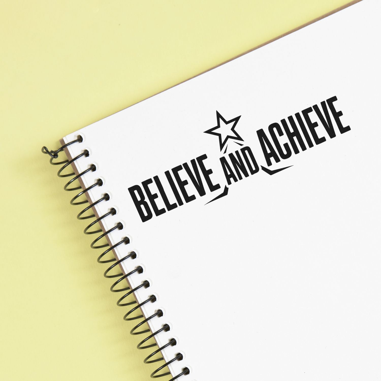 Black Believe and Achieve stamp/seal with star on a white spiral notebook, set against a pale yellow background.