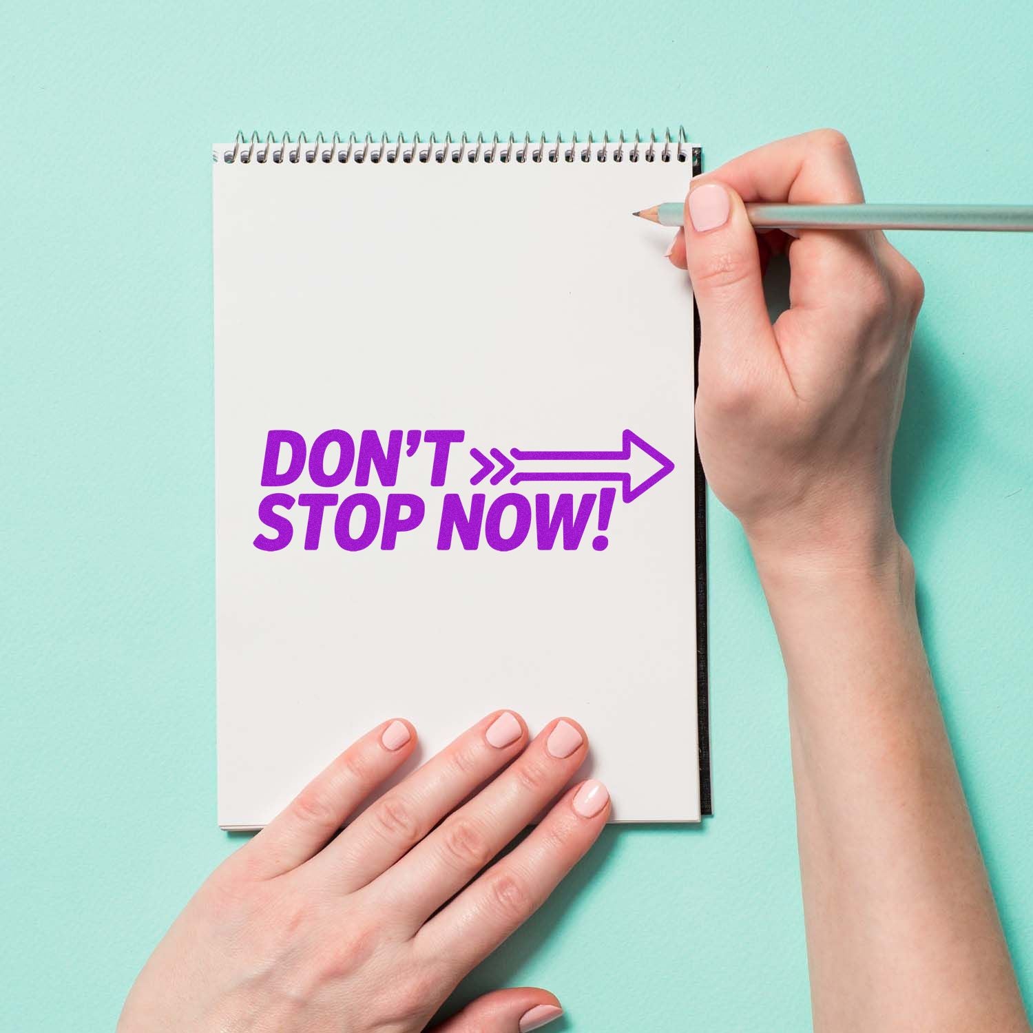 Purple DONT STOP NOW! stamp/seal on a white spiral notebook, with hand holding pencil on a mint background.