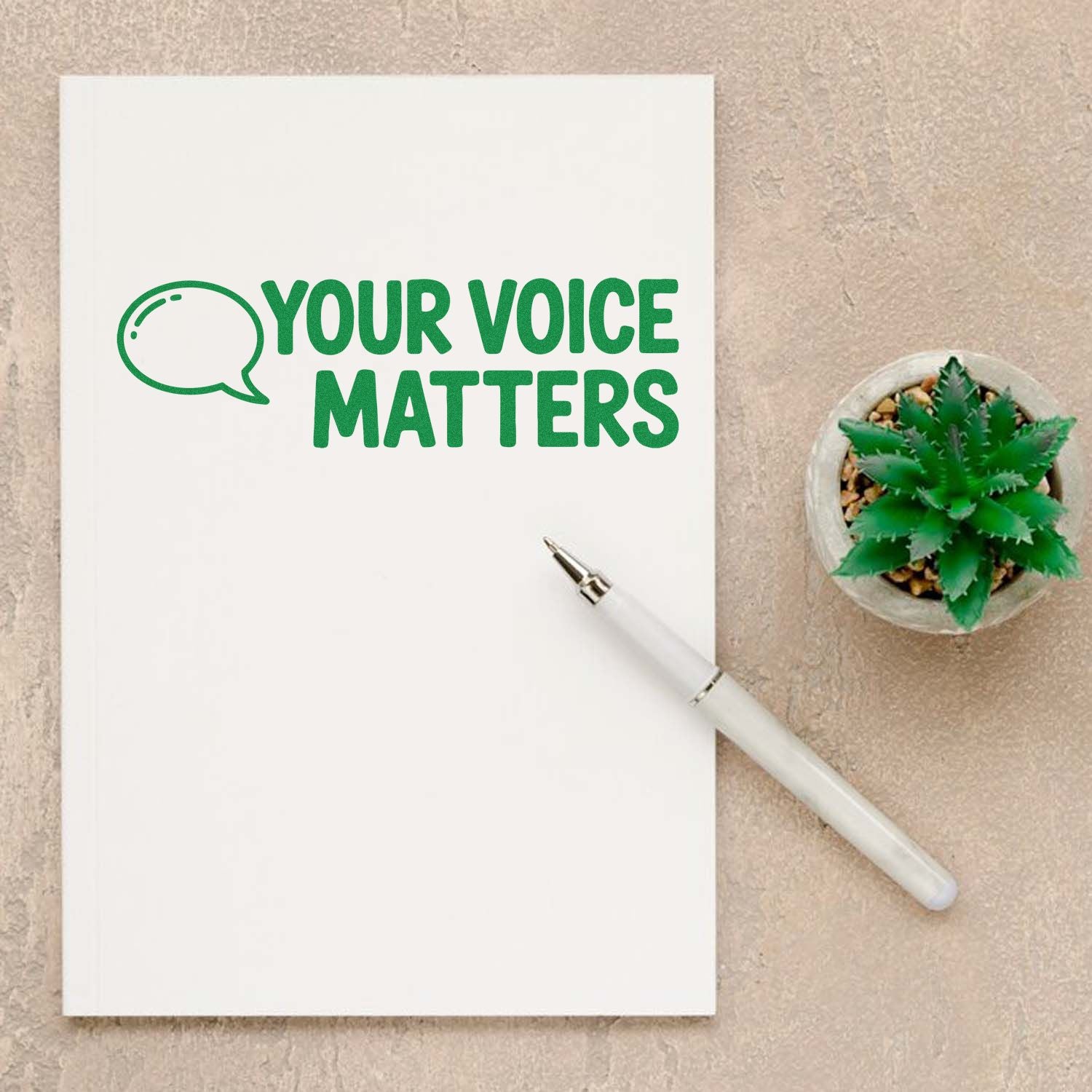 Green Your Voice Matters custom stamp on white notebook, with speech bubble icon, white pen, and potted succulent seal.