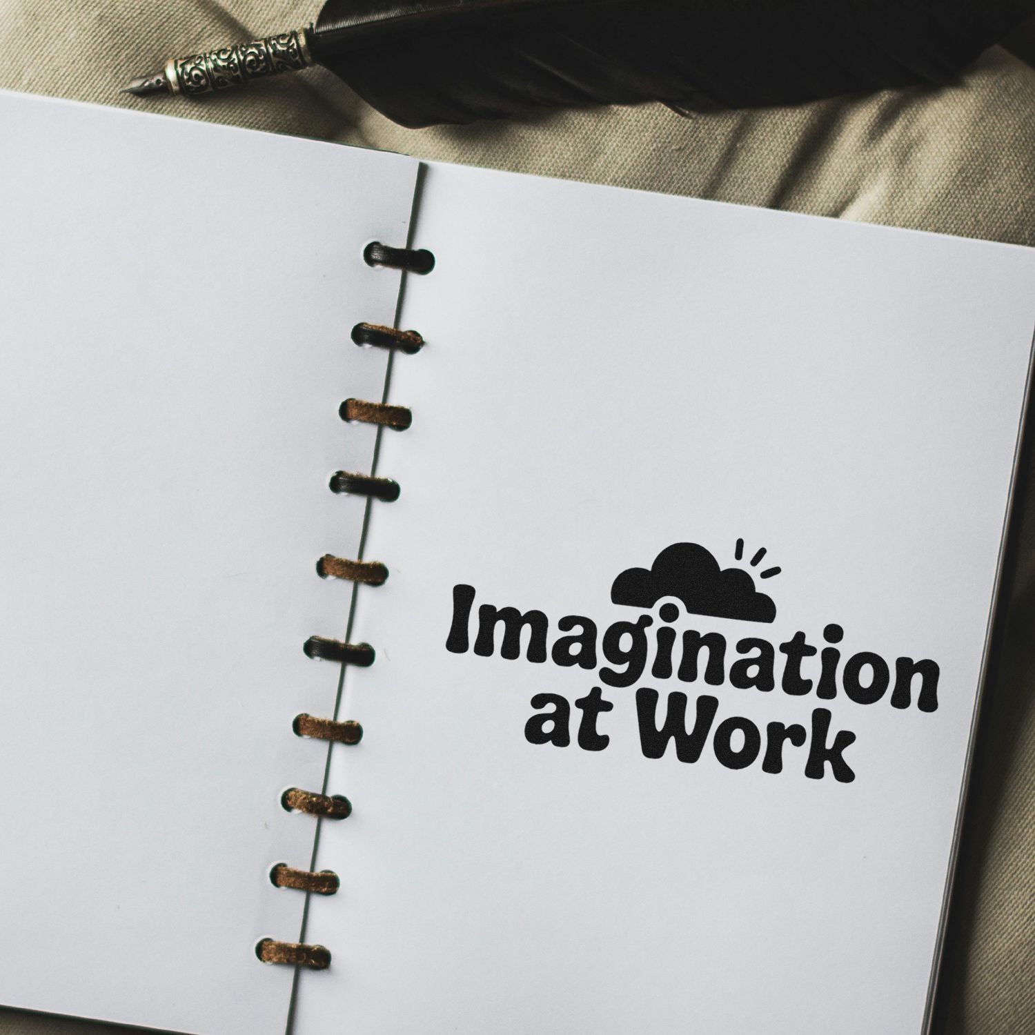 Black stamp and seal with Imagination at Work on white notebook page, spiral bound, beside a vintage pen.
