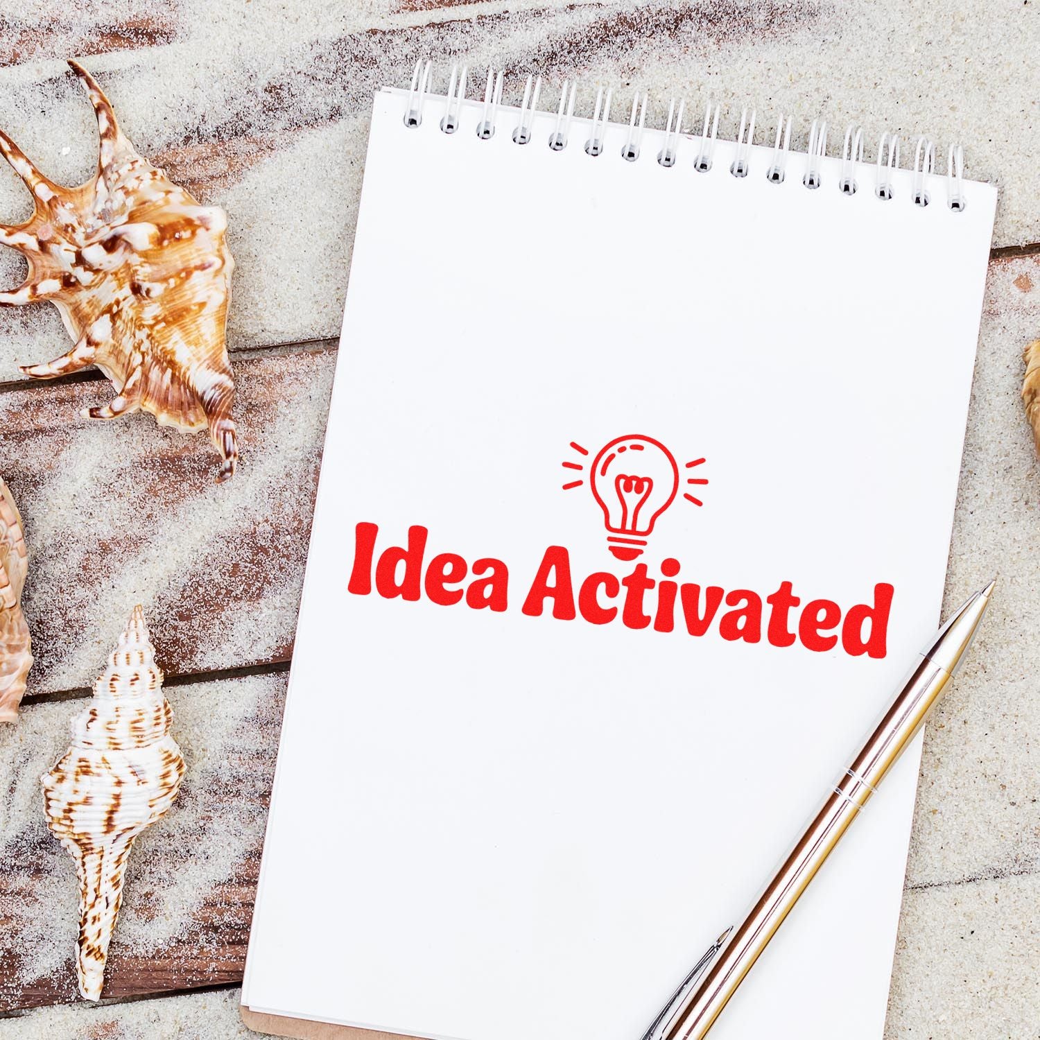 Red Idea Activated stamp on a white spiral notebook, with a lightbulb icon, seashells, and gold pen on sand.