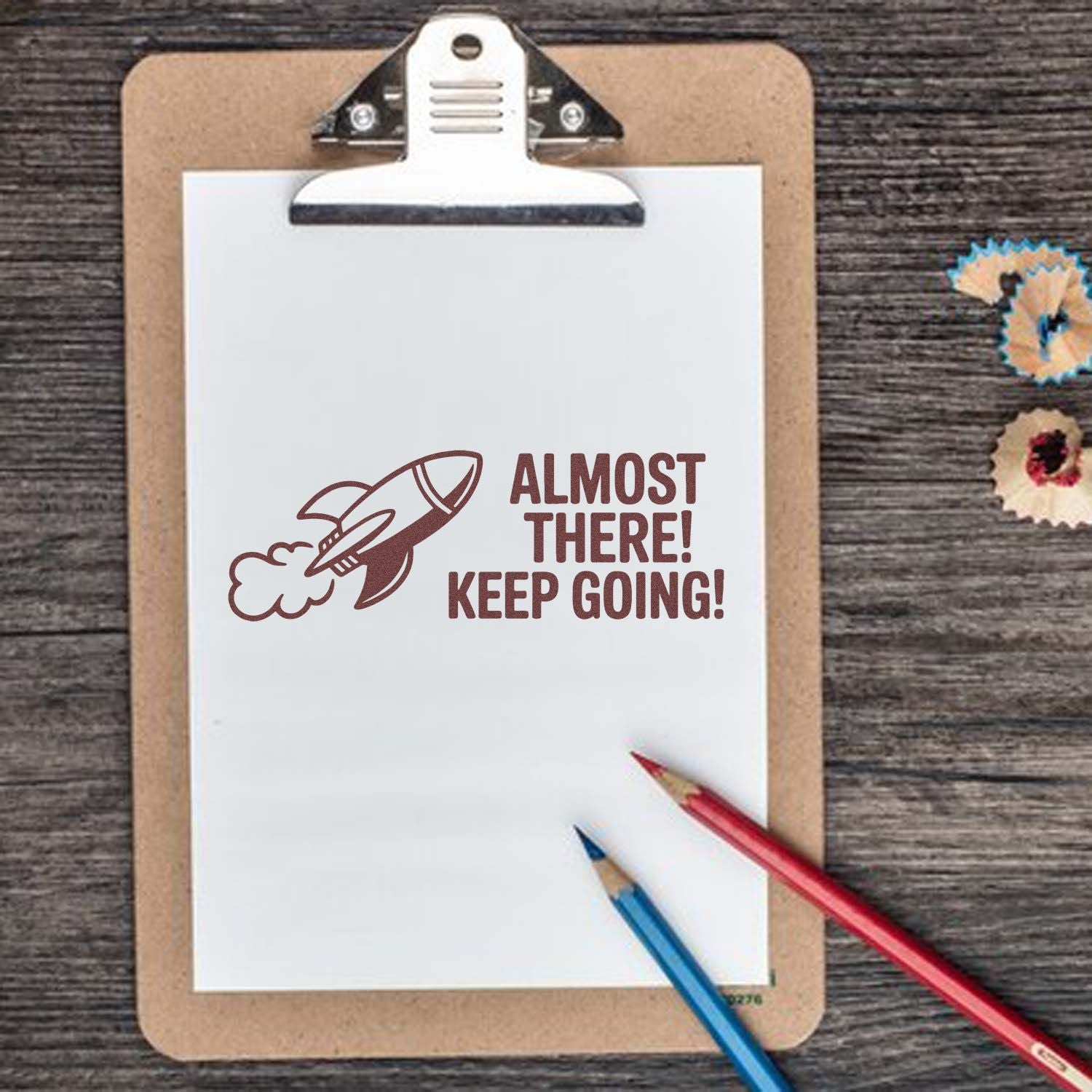 Clipboard with white paper, brown rocket stamp and Almost there! Keep going! text, plus red and blue pencils on rustic wood.