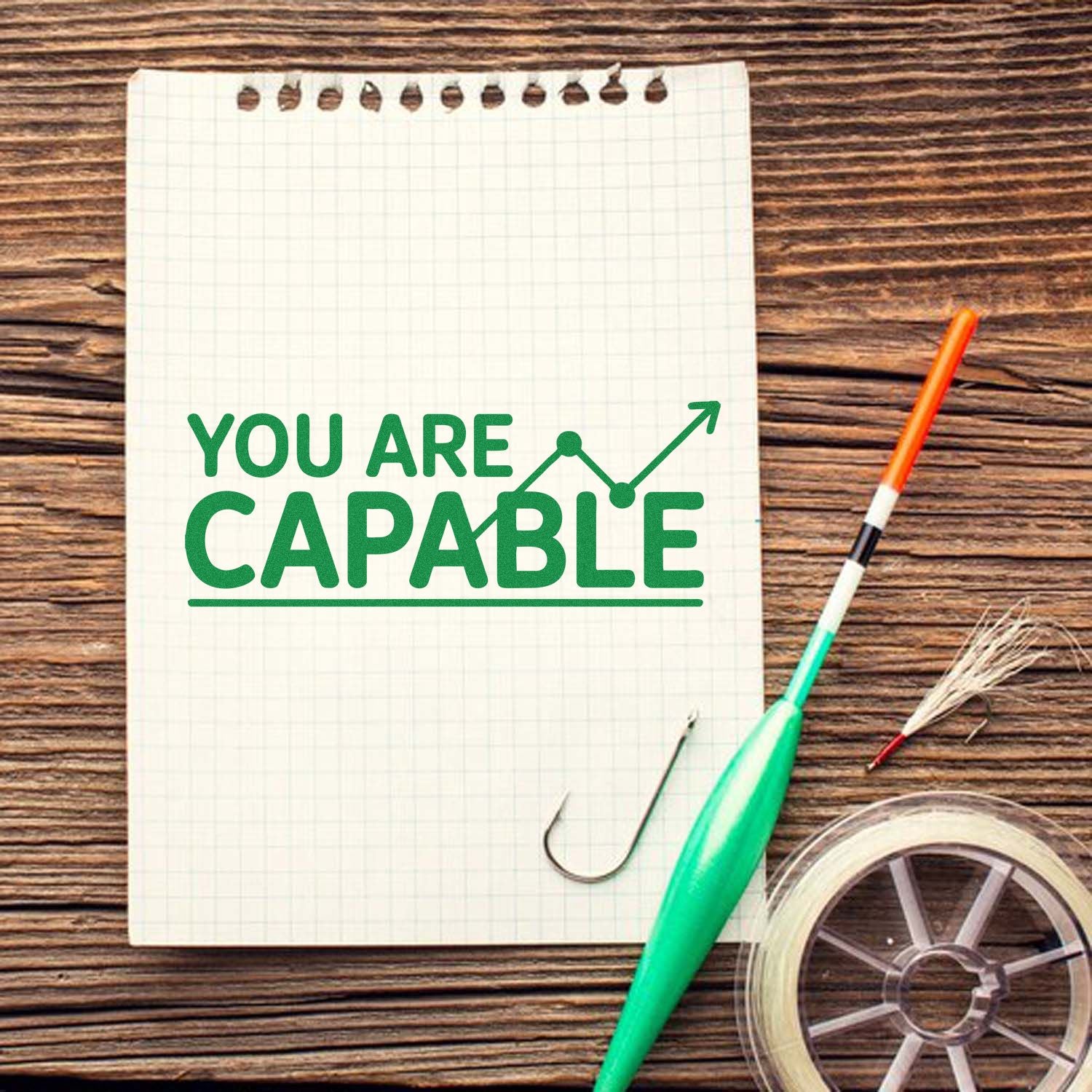 Green YOU ARE CAPABLE stamp-style text on a graph notebook, with arrow graphic, beside seal tape and a pen on wood.