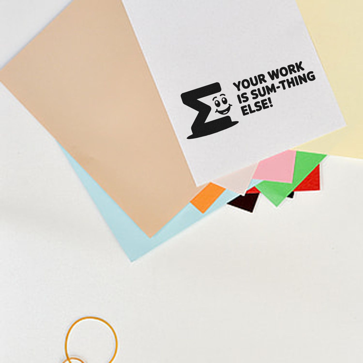Black self-inking stamp and seal on white paper, reading Your work is sum-thing else! with pastel colored sheets.