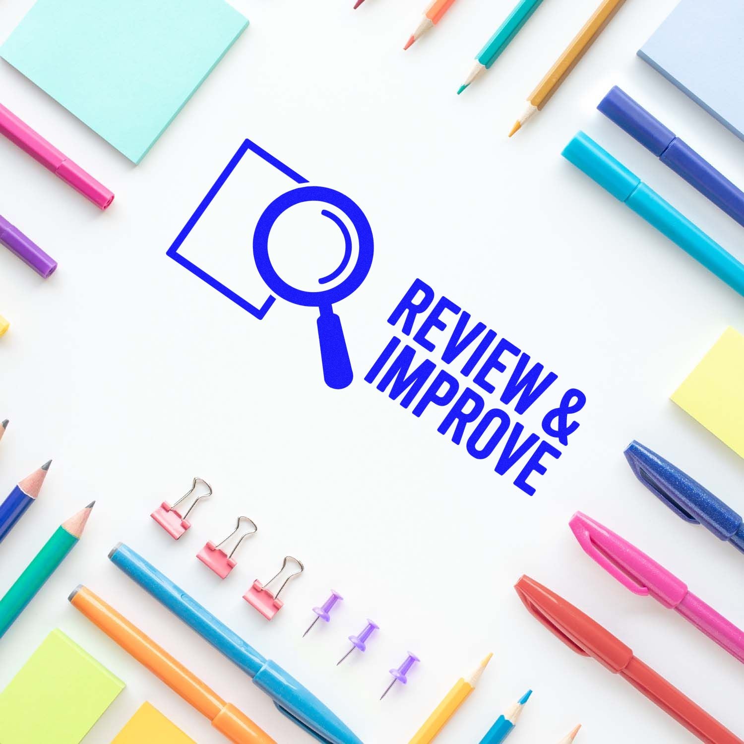 Blue review and improve stamp seal with magnifying glass, surrounded by colorful pens, pencils, binder clips, and sticky notes.