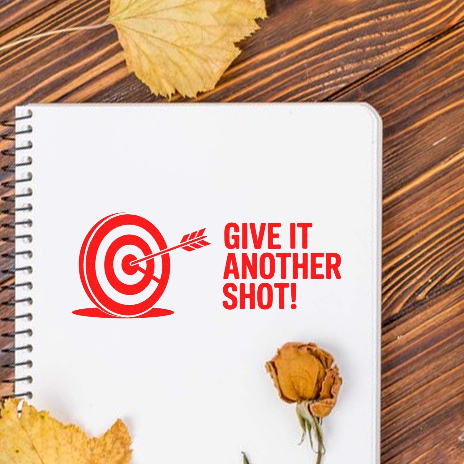 Red stamp-style target seal with arrow and GIVE IT ANOTHER SHOT! on a white notebook, on rustic wood with dried leaves.