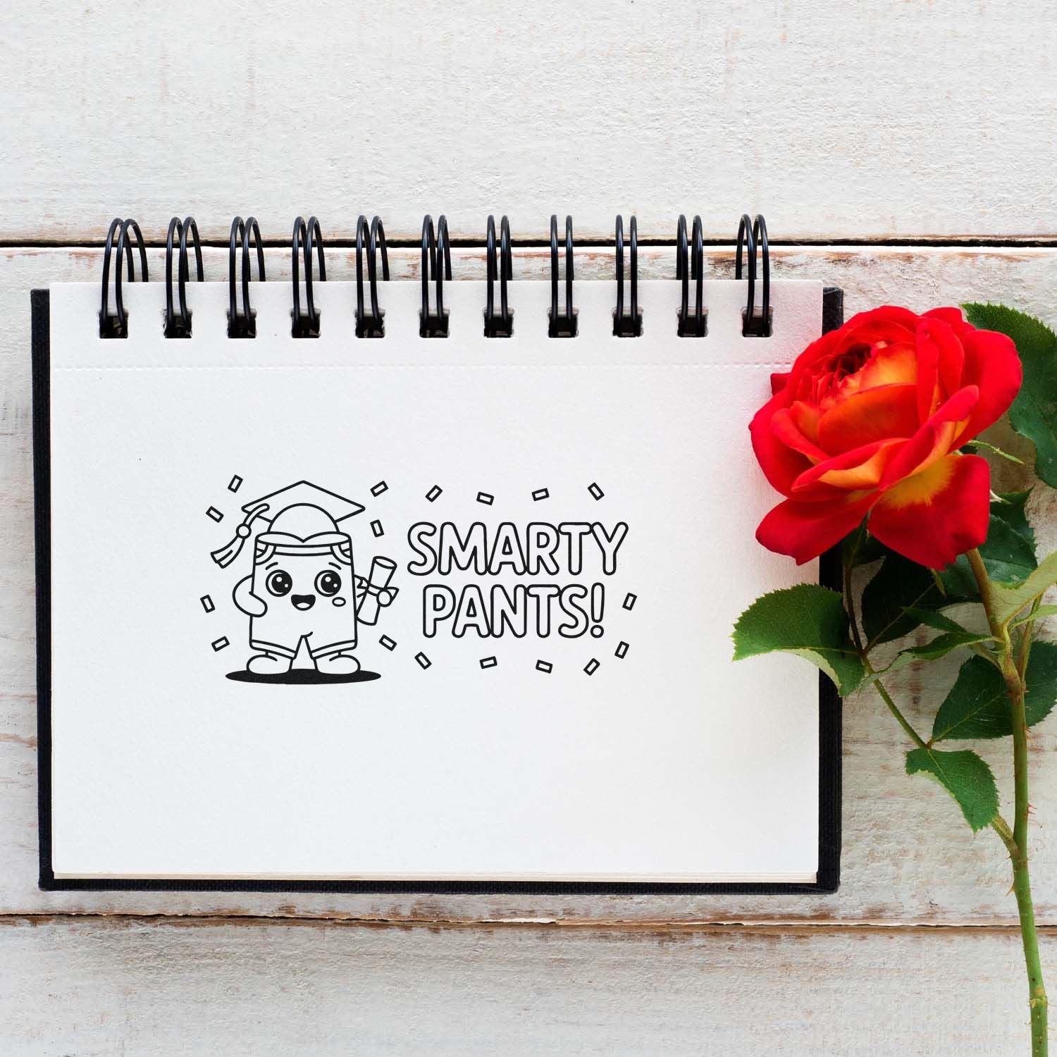 White spiral notebook with a black Smarty Pants graduation stamp and a red rose on a rustic wood background.