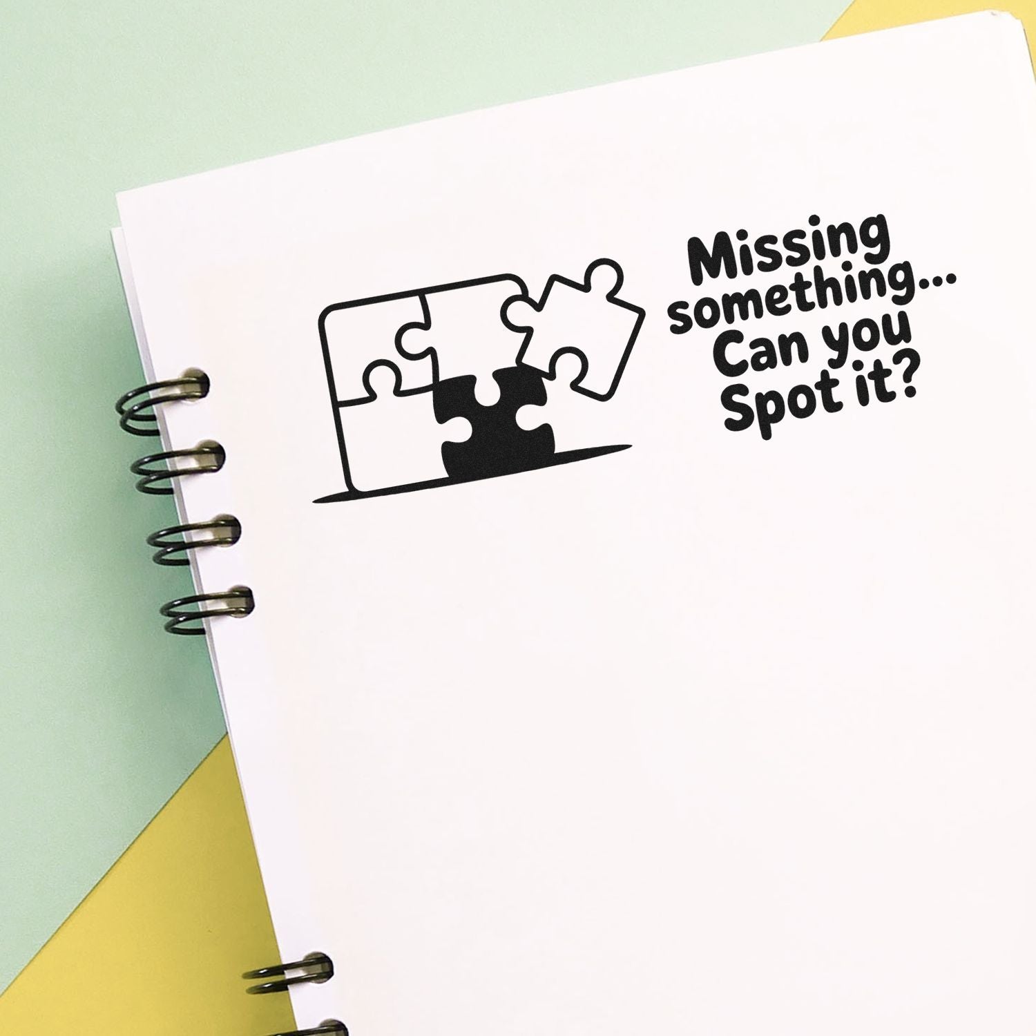 Black puzzle-piece stamp and seal reading Missing something... Can you spot it? on a white spiral notebook, mint and yellow background
