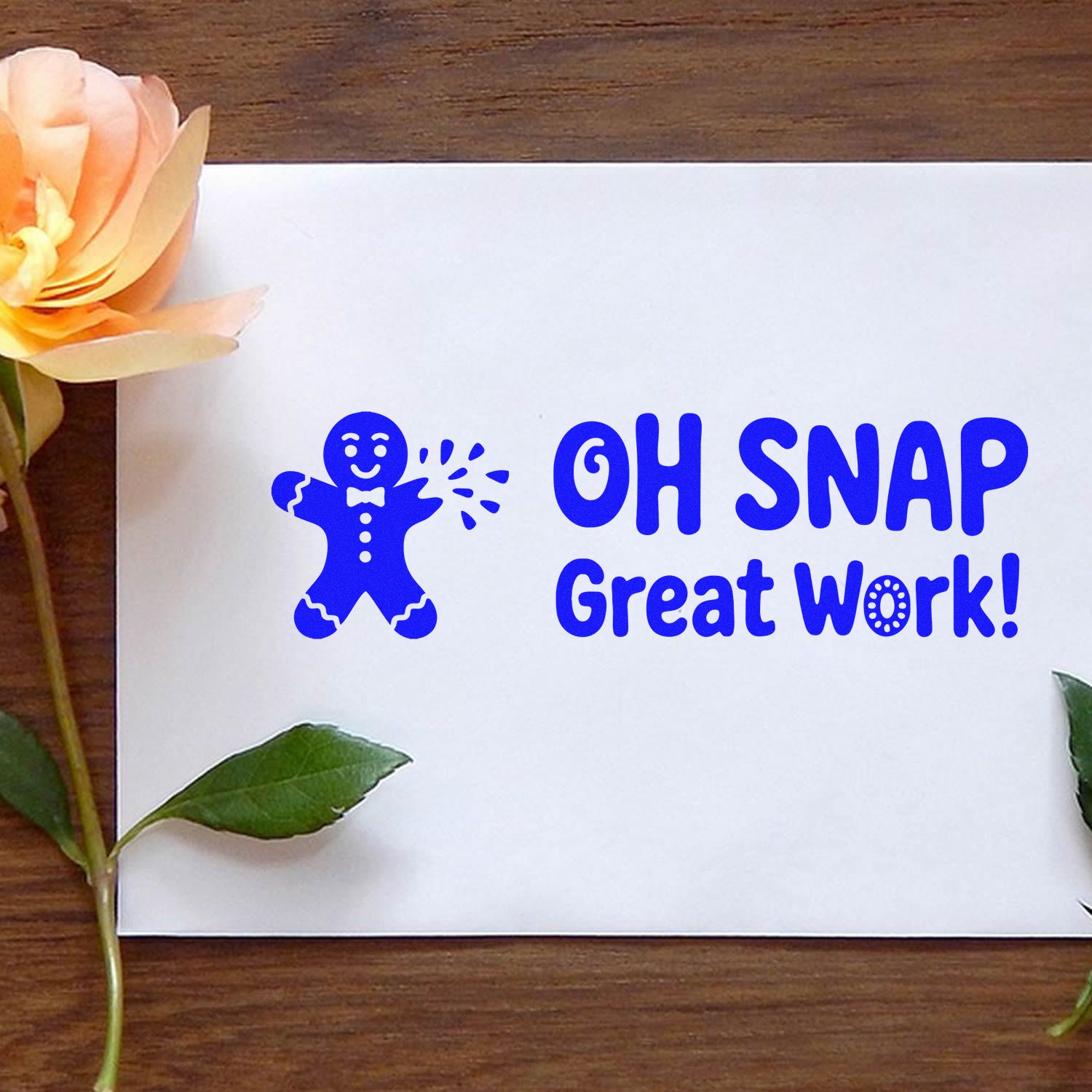 Blue stamp seal with Oh Snap Great Work! and gingerbread figure on a white card, with flowers on a wooden table.