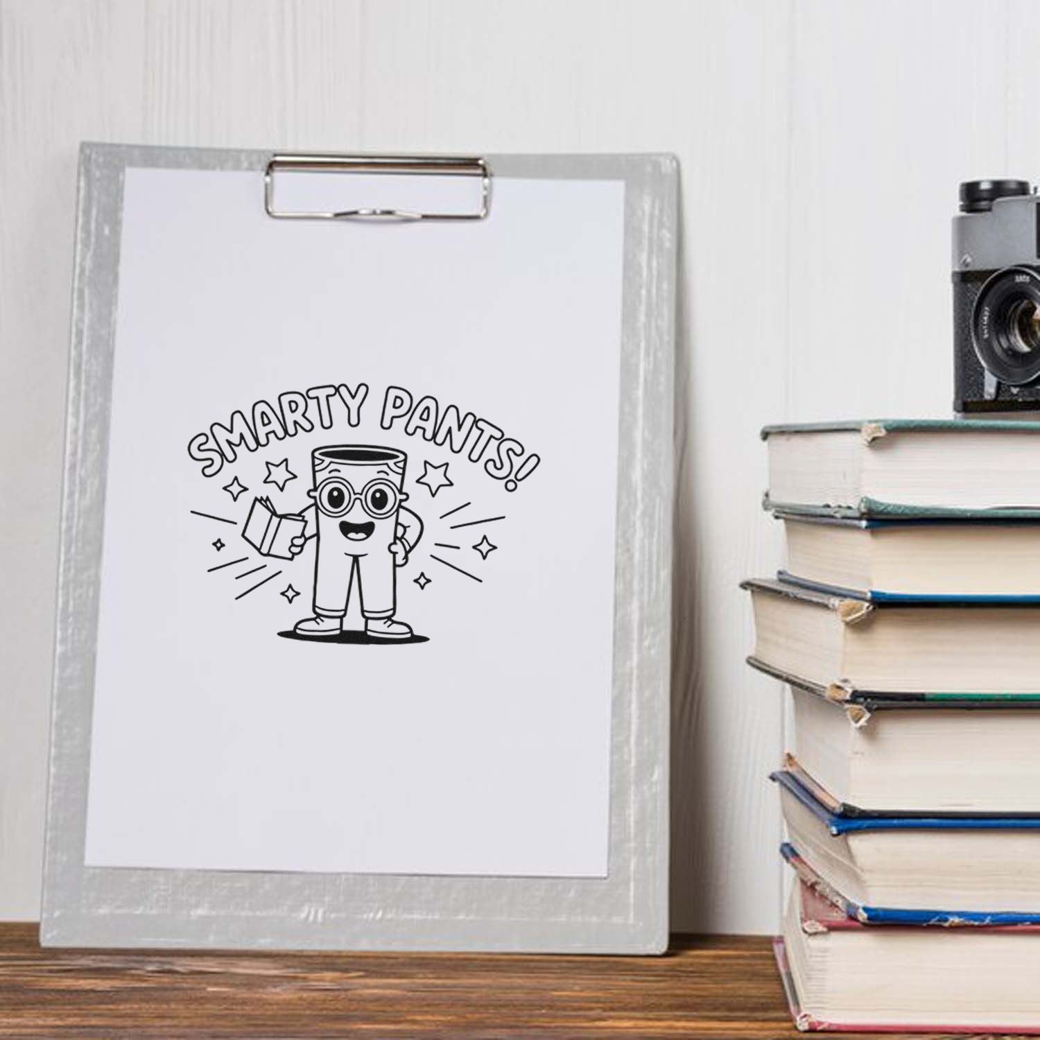 Black Smarty Pants stamp design on a white sheet clipped to a gray clipboard, beside stacked books and a vintage camera.