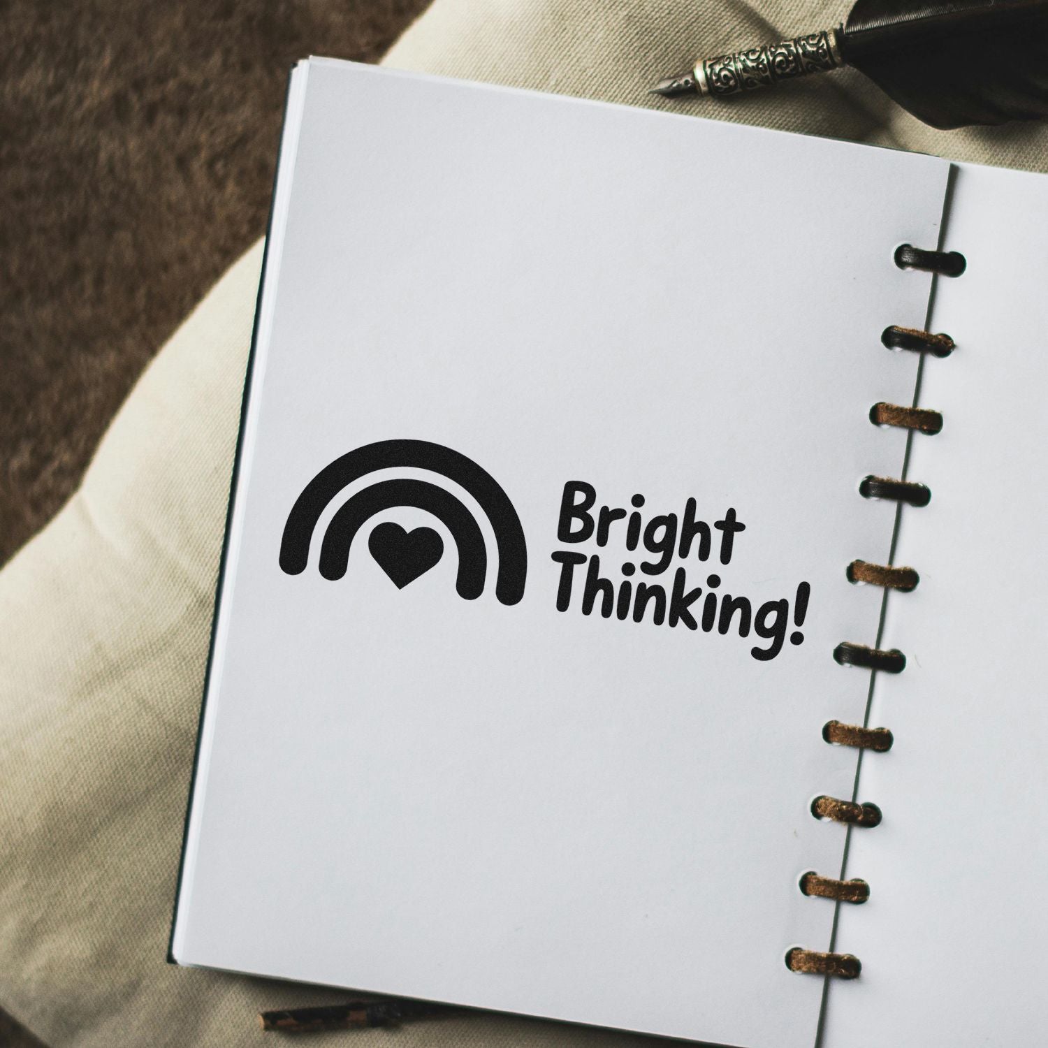Black stamp seal with rainbow-heart logo and Bright Thinking! on a white spiral-bound notebook page, bronze rings visible.