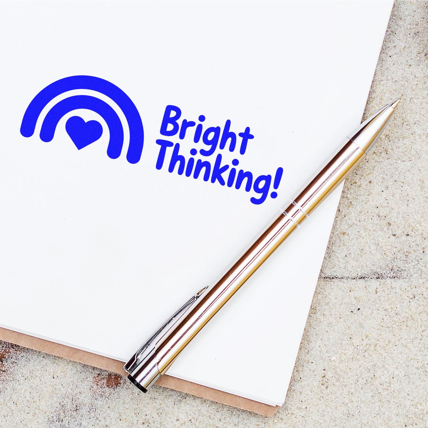 Blue Bright Thinking! stamp seal with rainbow heart logo on white paper, beside a silver and gold pen.