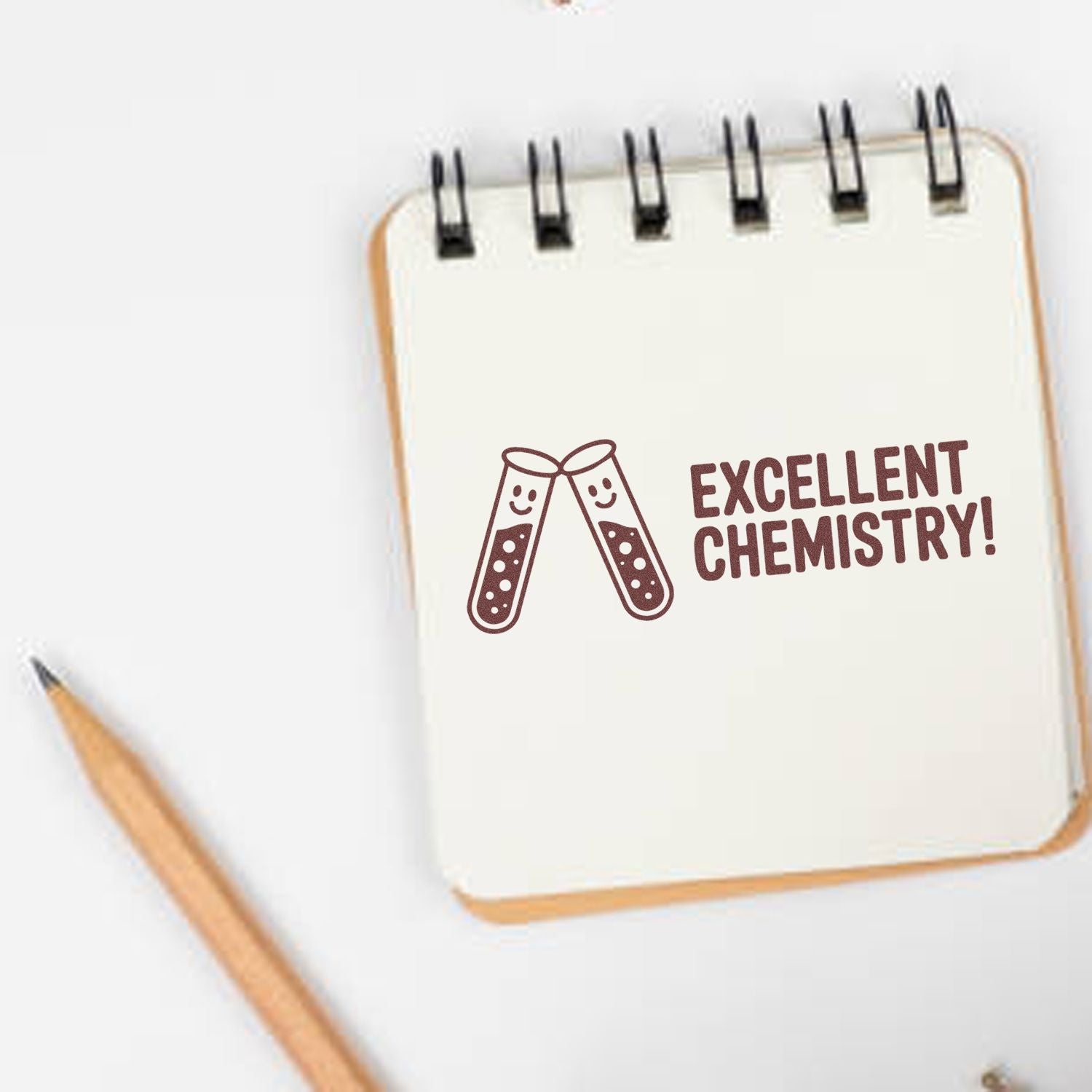 Brown chemistry stamp seal with smiling test tubes and Excellent Chemistry! on a white spiral notebook beside a pencil.