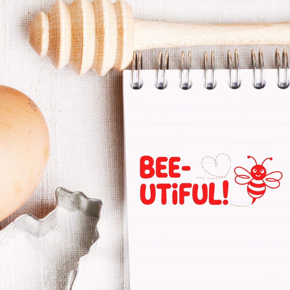 Red bee-themed stamp on a spiral notebook, with wooden seal tools, egg, and metal cookie cutter on beige fabric.