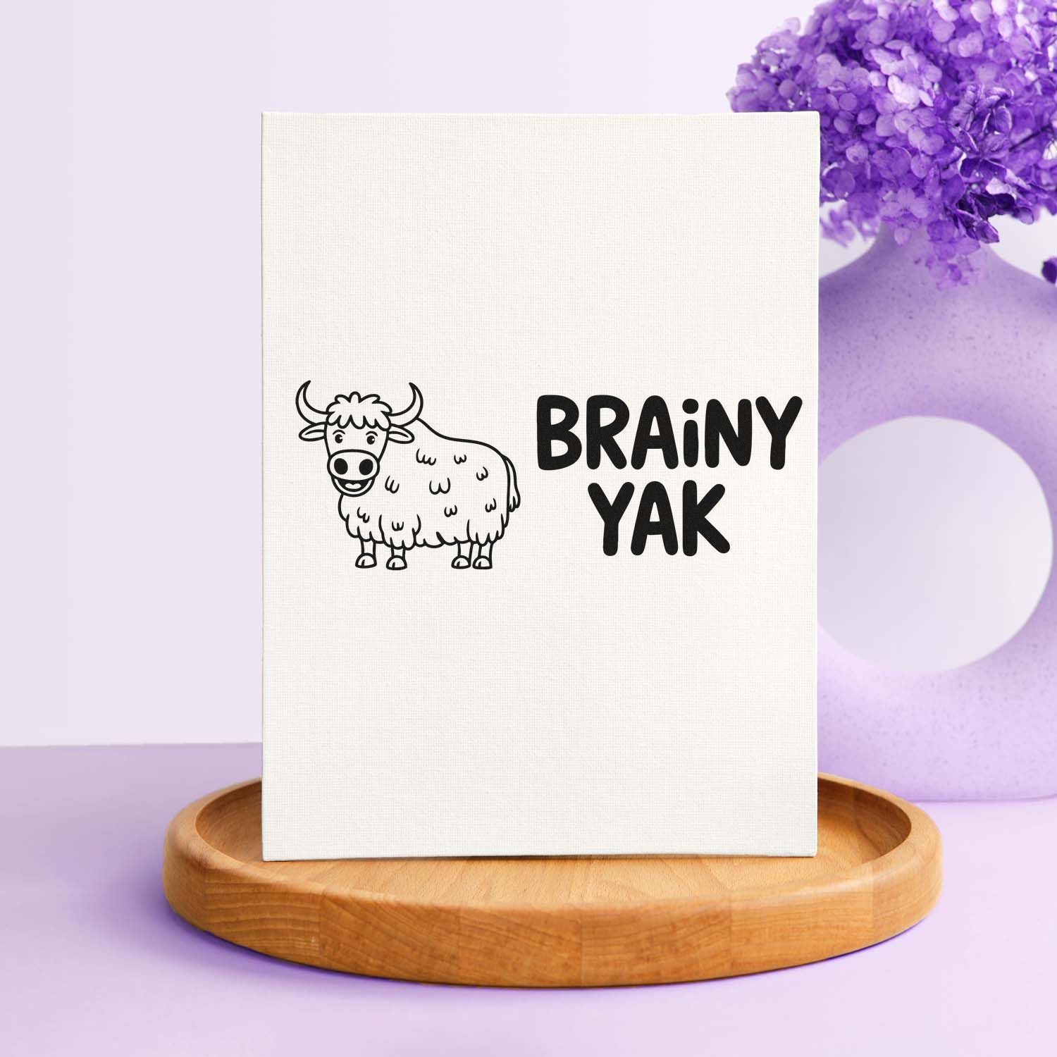 White custom stamp seal with black cartoon yak and BRAINY YAK text, on a lavender background with purple florals and a wooden base