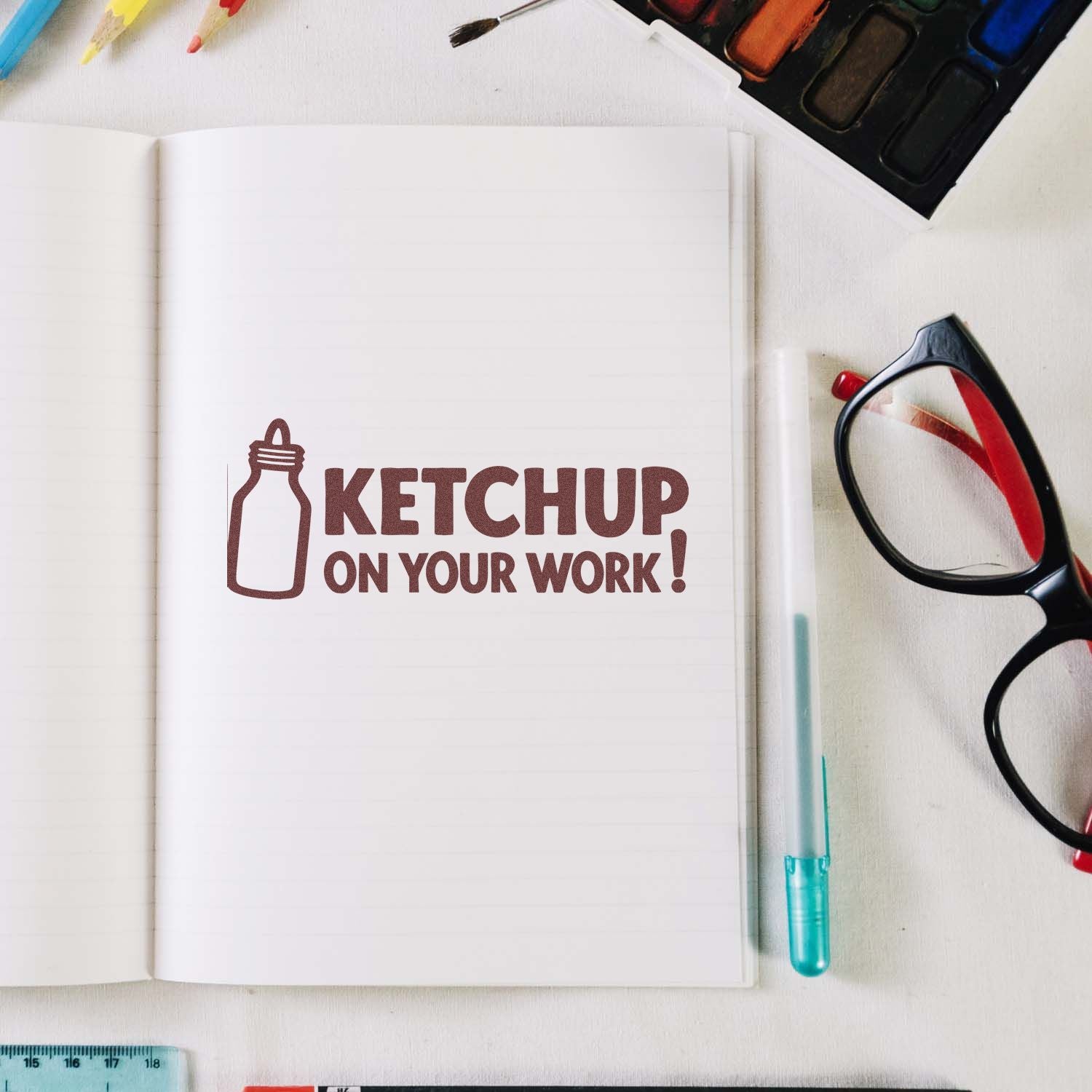 Brown stamp and seal reading KETCHUP ON YOUR WORK! on a white notebook, with bottle icon and art supplies.
