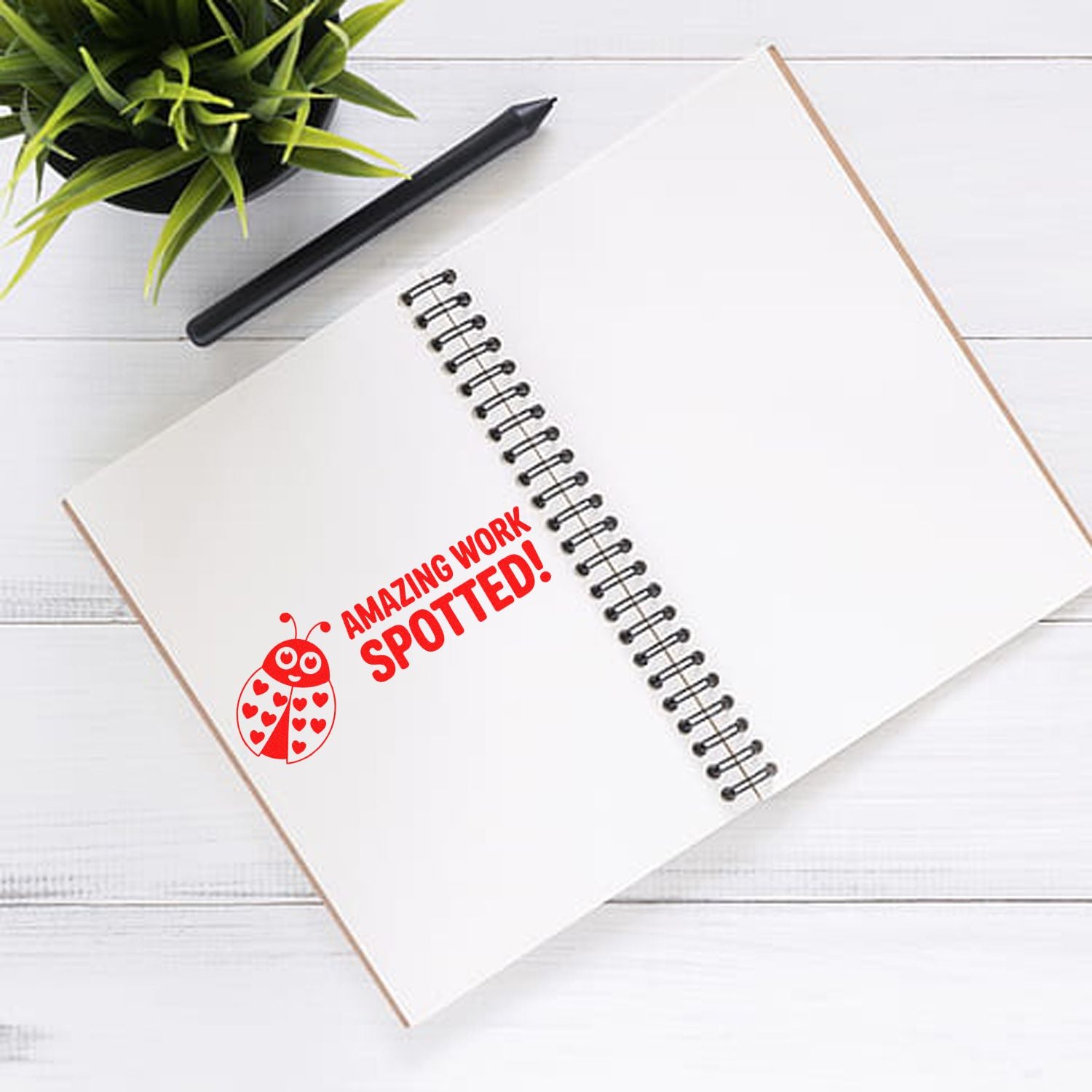 Red self-inking ladybug stamp reading Amazing Work Spotted! on a white spiral notebook with black pen and green plant