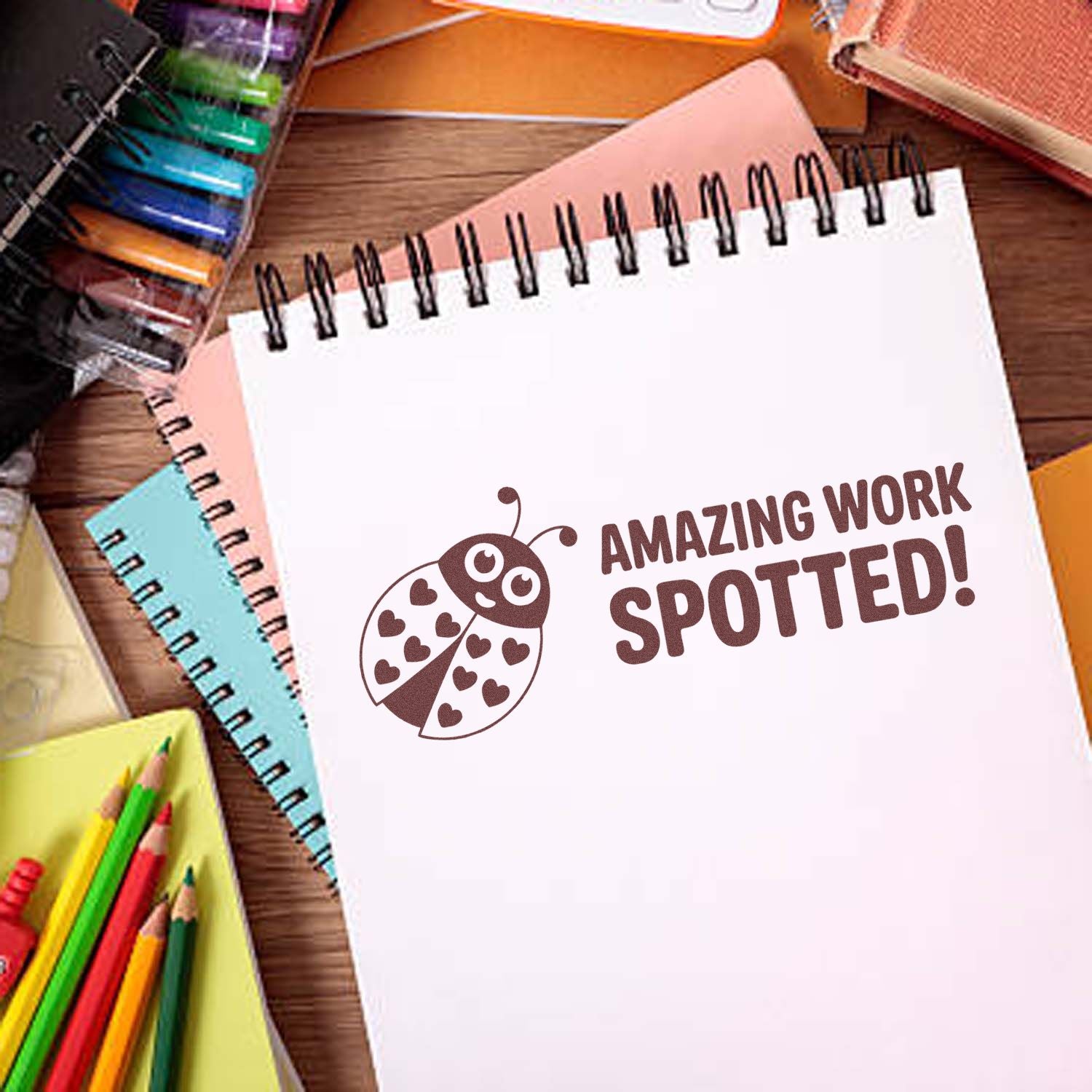 Brown ladybug stamp seal reading AMAZING WORK SPOTTED! on a white spiral notebook, surrounded by colorful stationery.