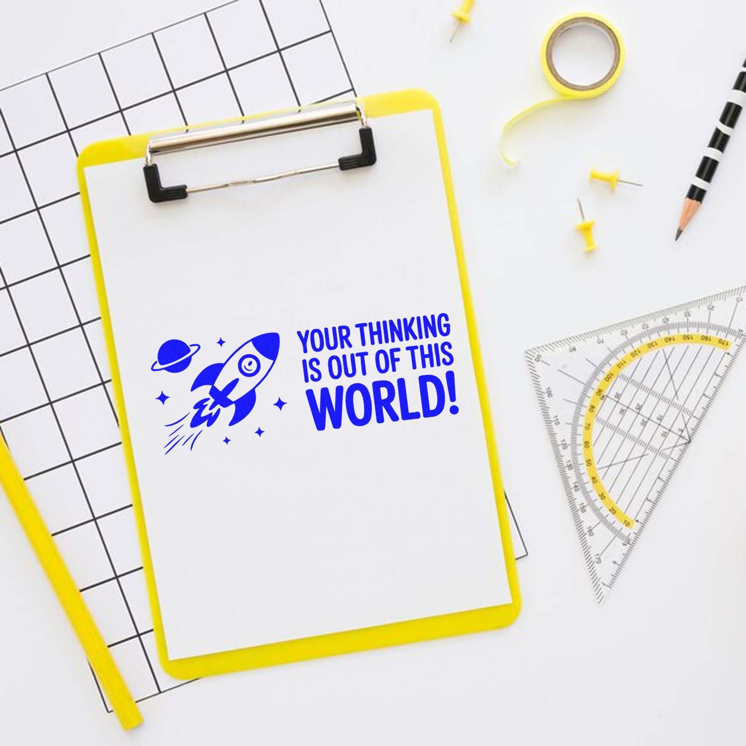 Yellow clipboard with blue rocket stamp and seal, Your thinking is out of this world! beside drafting tools.