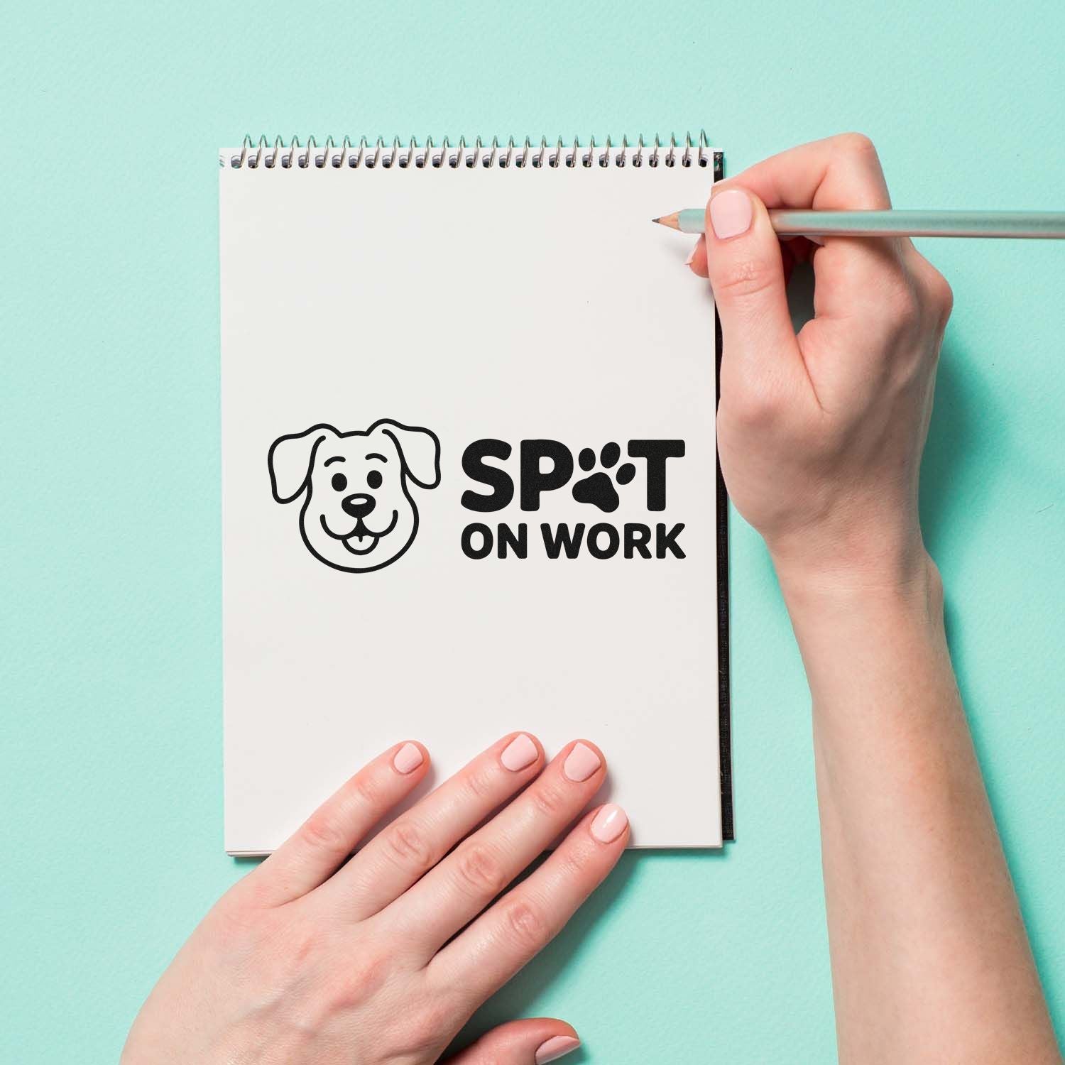 Black dog logo stamp and seal reading SPOT ON WORK on a white spiral notebook, with a hand holding a pencil on mint green.