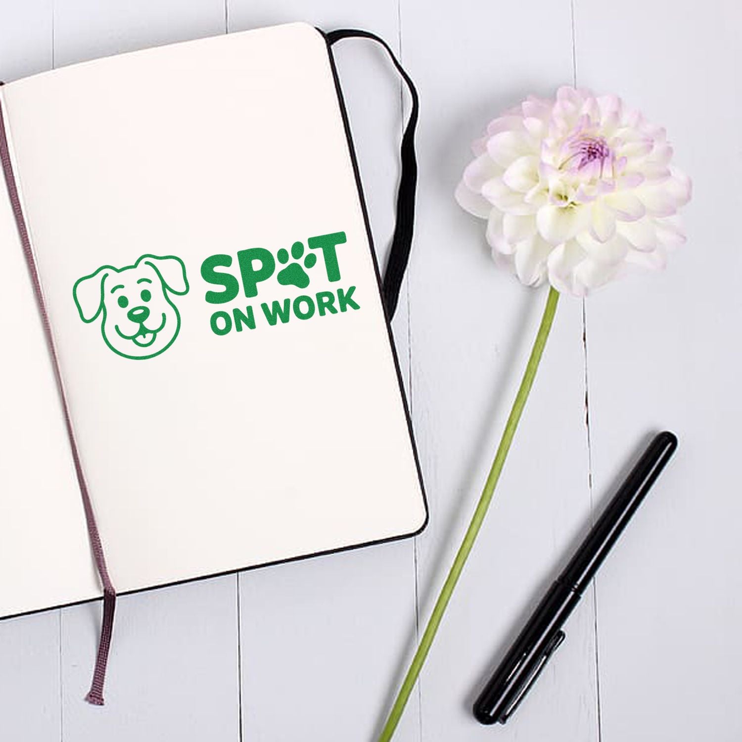 Green dog-logo stamp seal reading SPOT ON WORK on a cream notebook, beside a white flower and black pen.