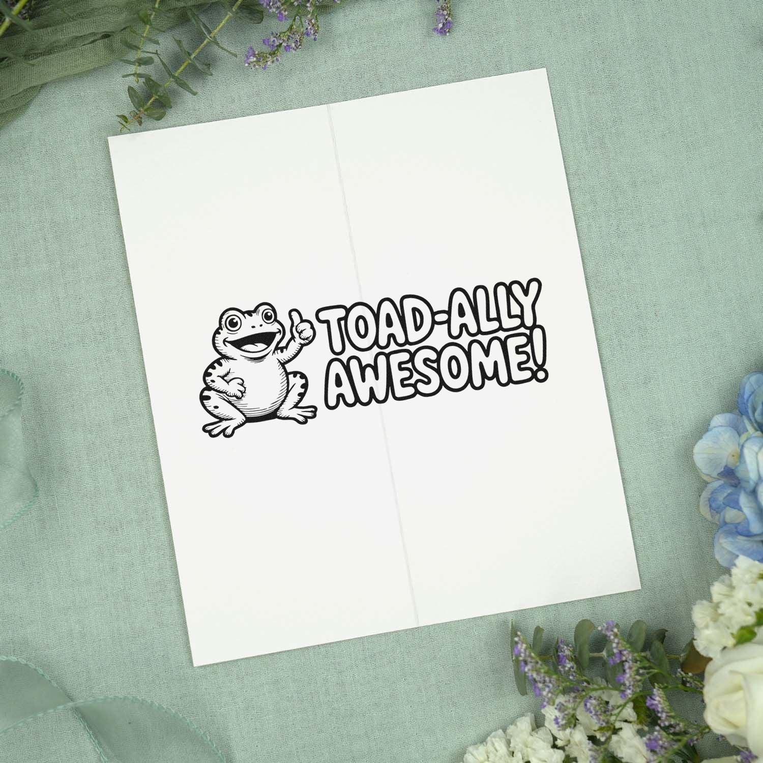 Black frog stamp and seal reading Toad-ally Awesome! on white card, set on mint fabric with purple flowers.