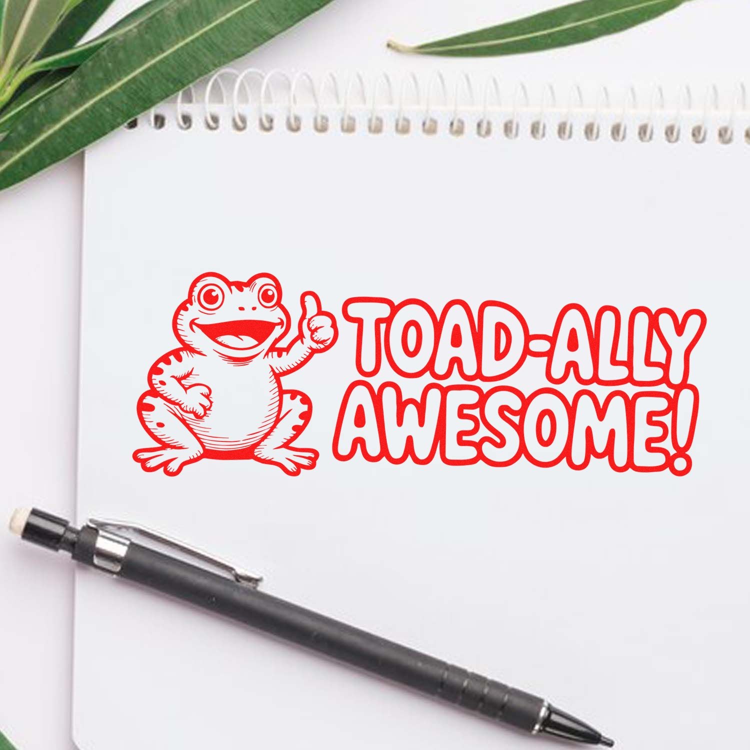 Red frog rubber stamp seal reading TOAD-ALLY AWESOME! on a white notebook, with green leaves and a black pen.