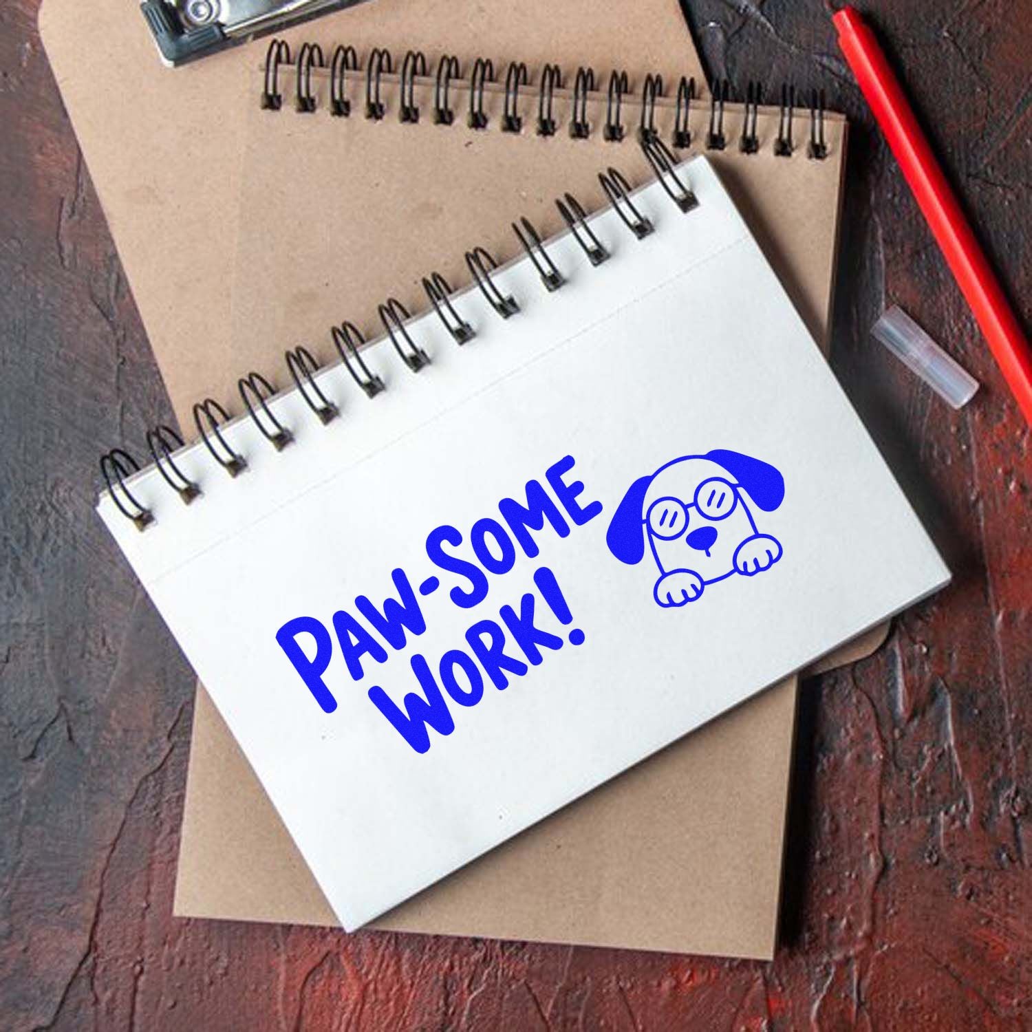 Blue dog stamp and seal on white spiral notebook reading Paw-some Work! with brown notebook and red pen.
