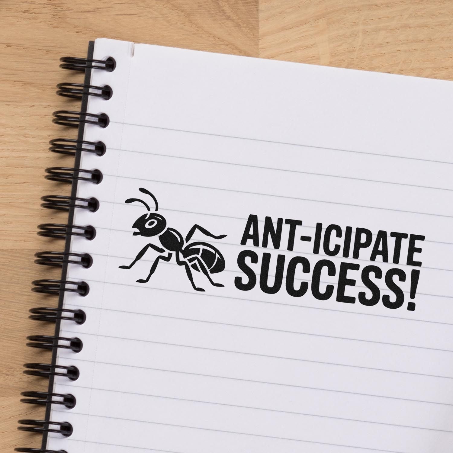 Black ant stamp seal on a white lined notebook reads ANT-ICIPATE SUCCESS! with black spiral binding.