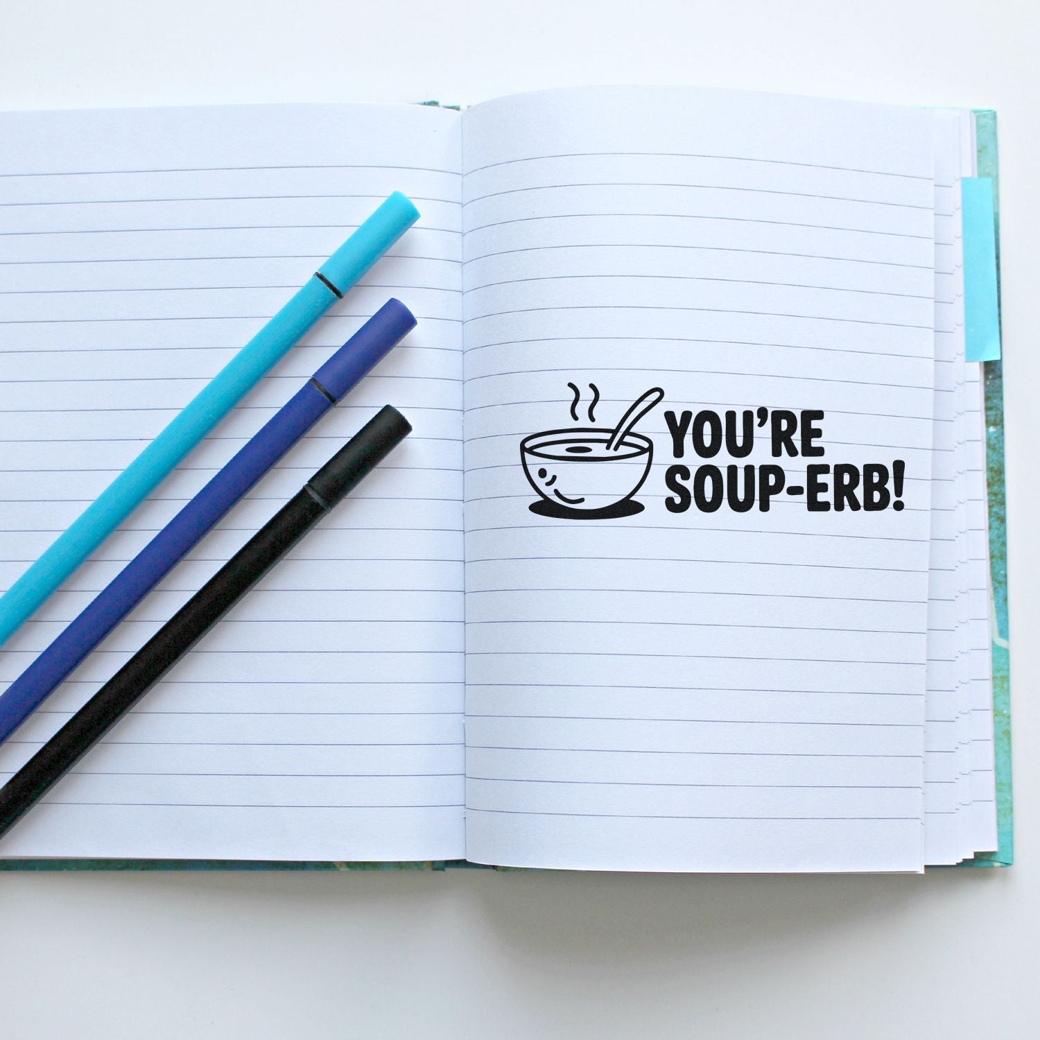 Open lined notebook with black soup stamp seal reading YOURE SOUP-ERB! and three turquoise, purple, and black pens.