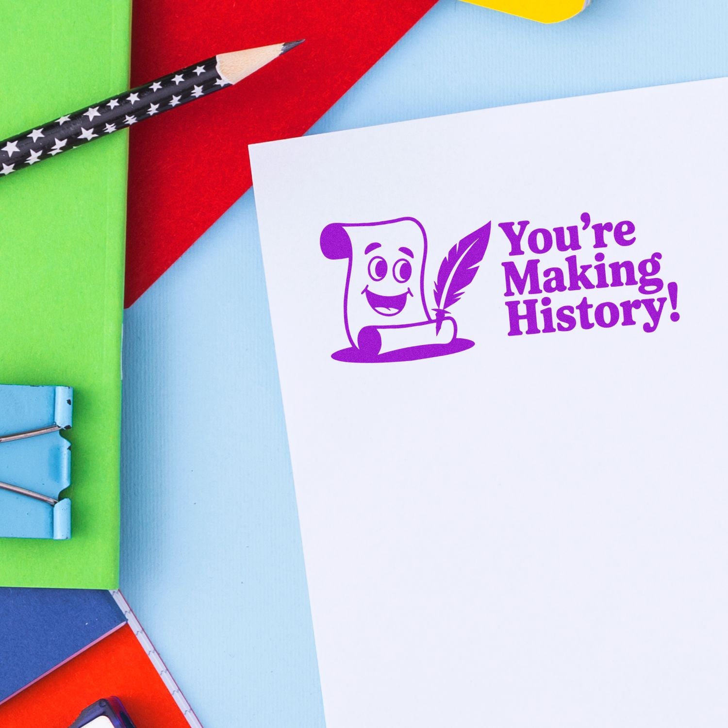 Purple rubber stamp seal reading Youre Making History! on white paper, with colorful pencils and bright paper background.