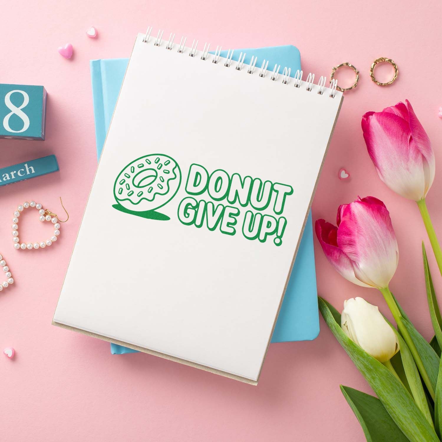 Green donut DONUT GIVE UP! stamp on a white spiral notebook, with pastel pink tulips and stationery accents.