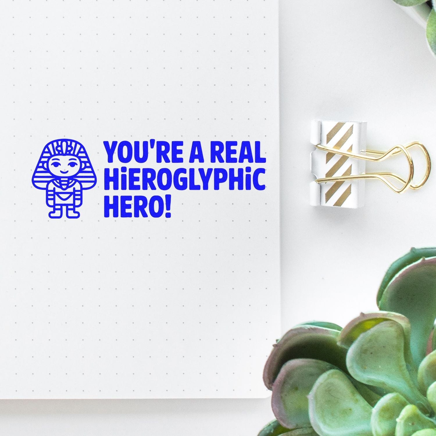 Blue Egyptian-themed stamp reading Youre a real hieroglyphic hero! on dotted white notebook, with gold-striped paperclip seal.